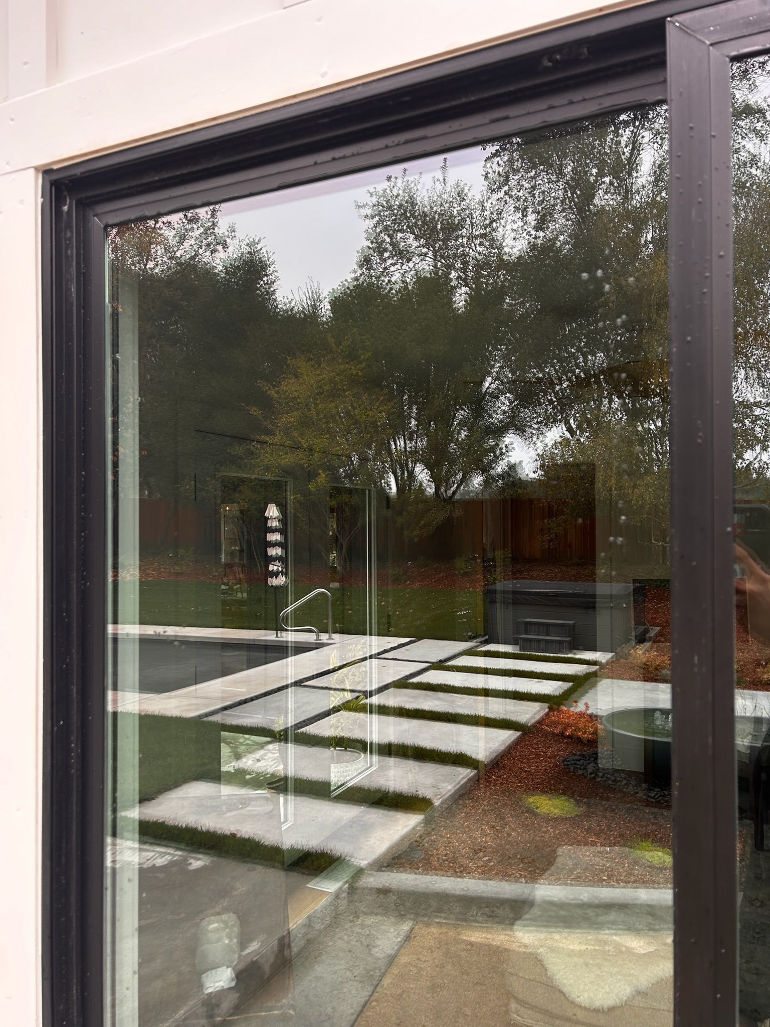 Black-framed window reflects an outdoor patio with a pool and trees on a cloudy day.