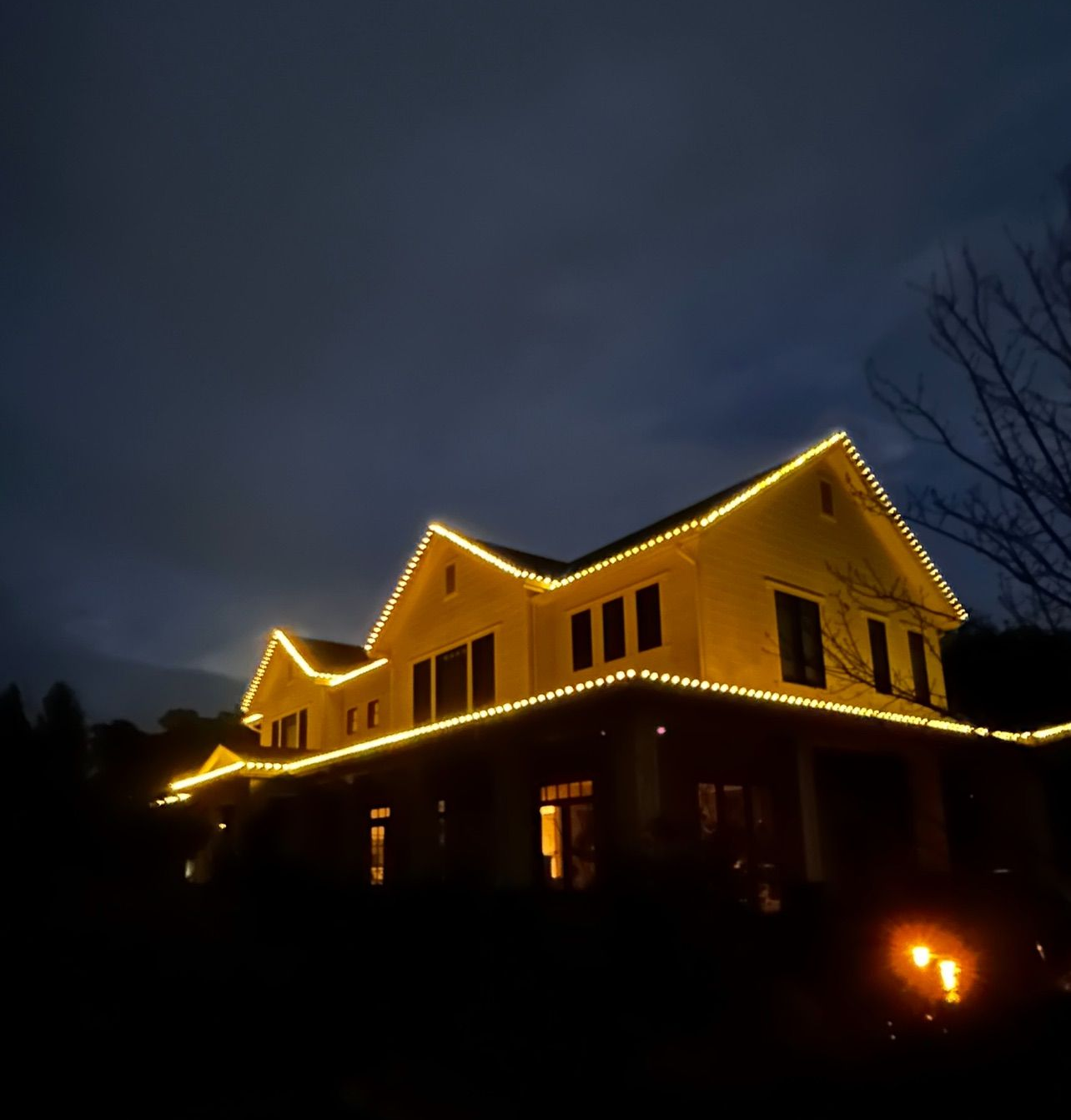 A house at dusk, lit with yellow lights along the roof and porch, against a dark blue sky.