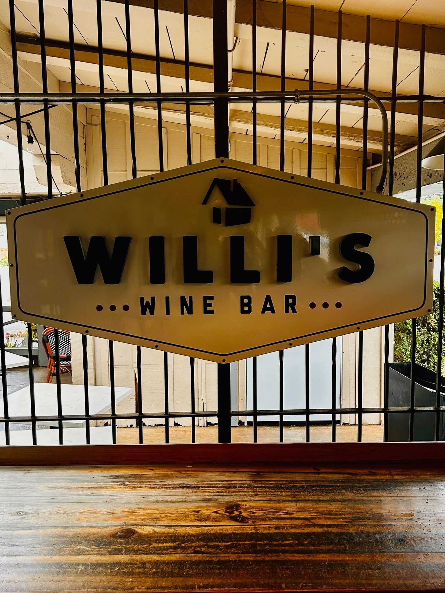 Sign for Willi's Wine Bar, black text on a cream-colored shape. Wooden bar in foreground, metal gate in background.