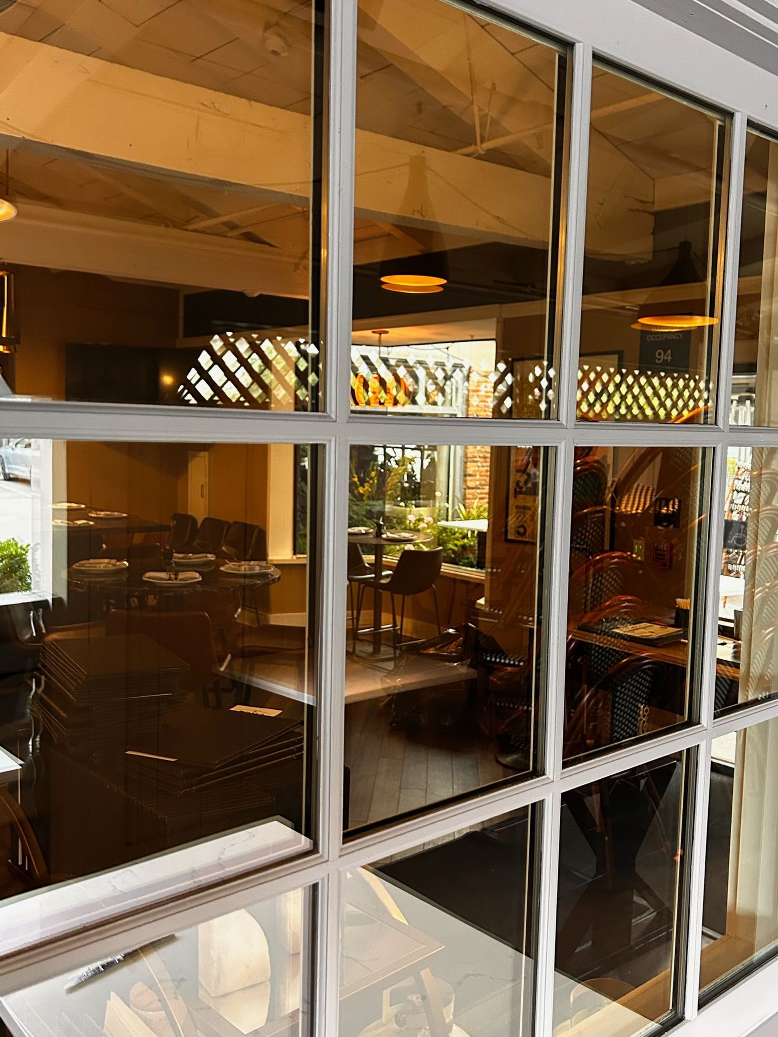 View through a multi-paned window of a restaurant interior. Tables and lighting are visible with an outdoor area.