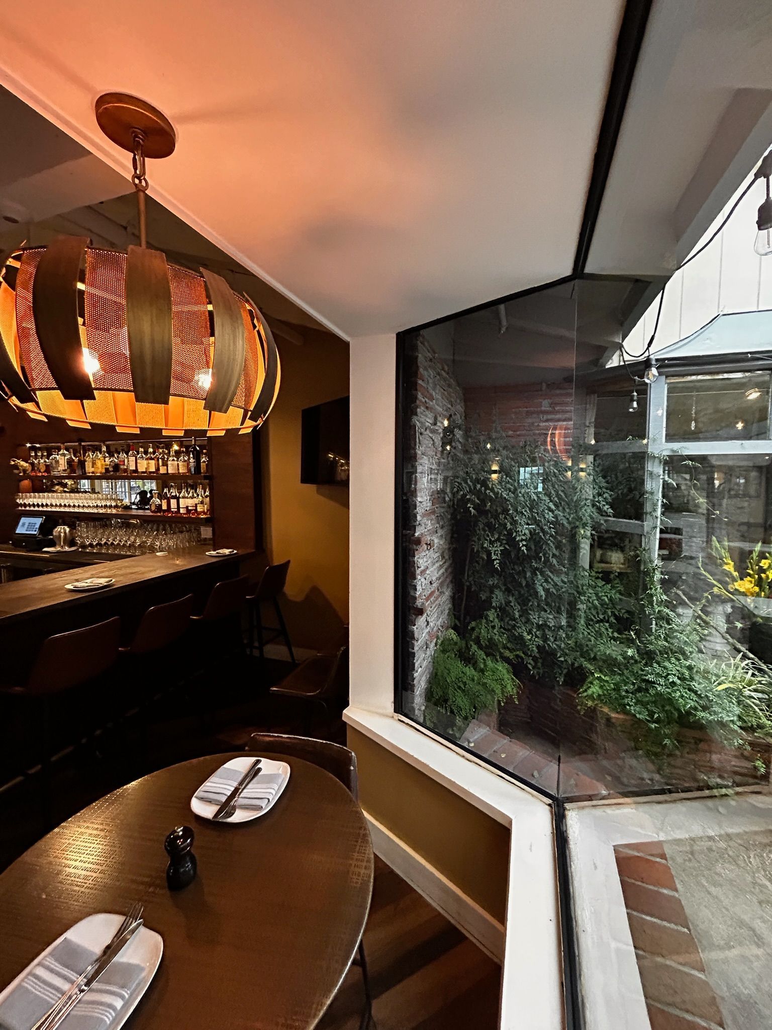 Restaurant interior with bar, table, and large window overlooking a small garden. Decorative light fixture.