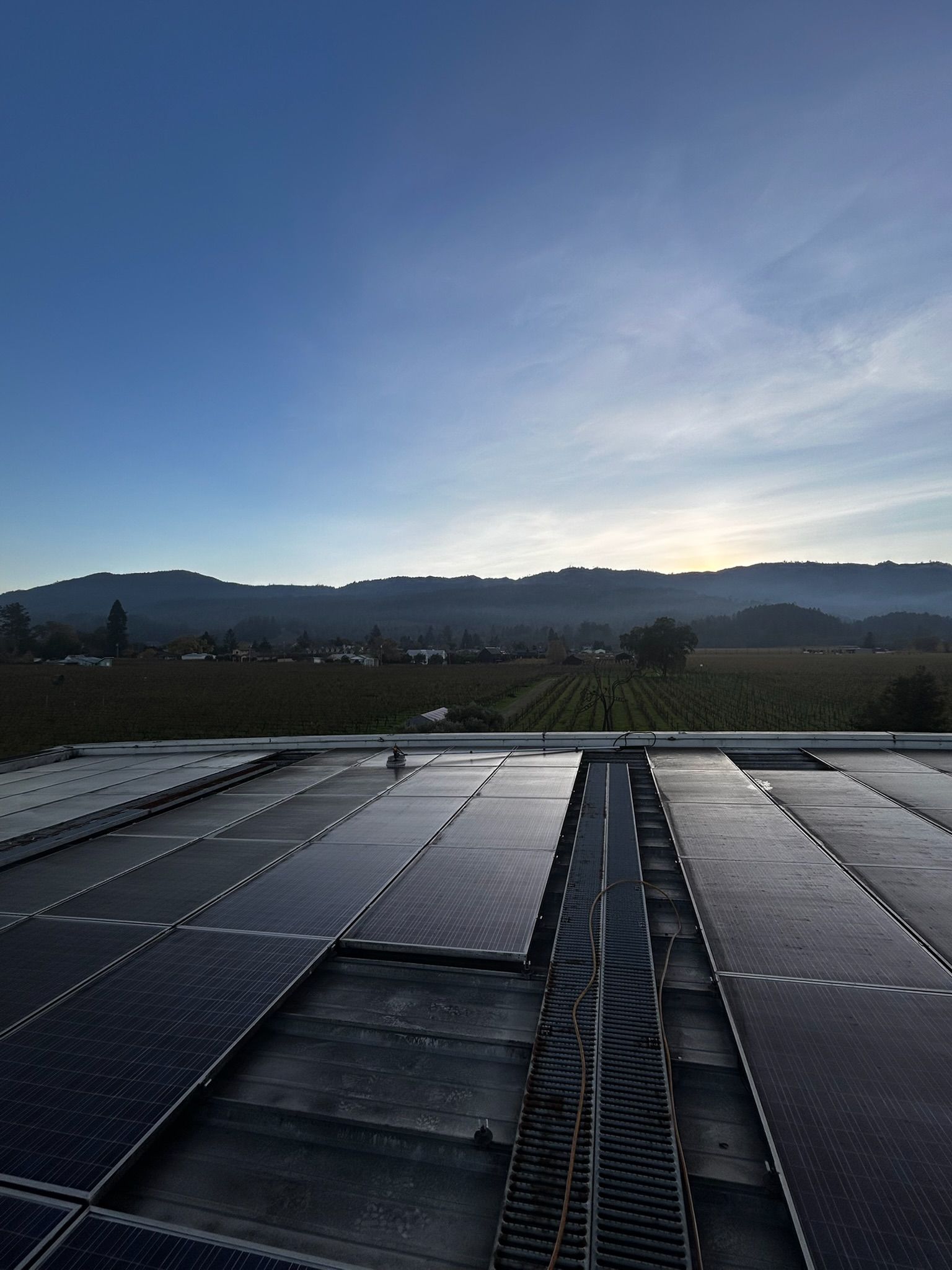 Solar panels on a roof with a landscape of fields and mountains under a blue sky.