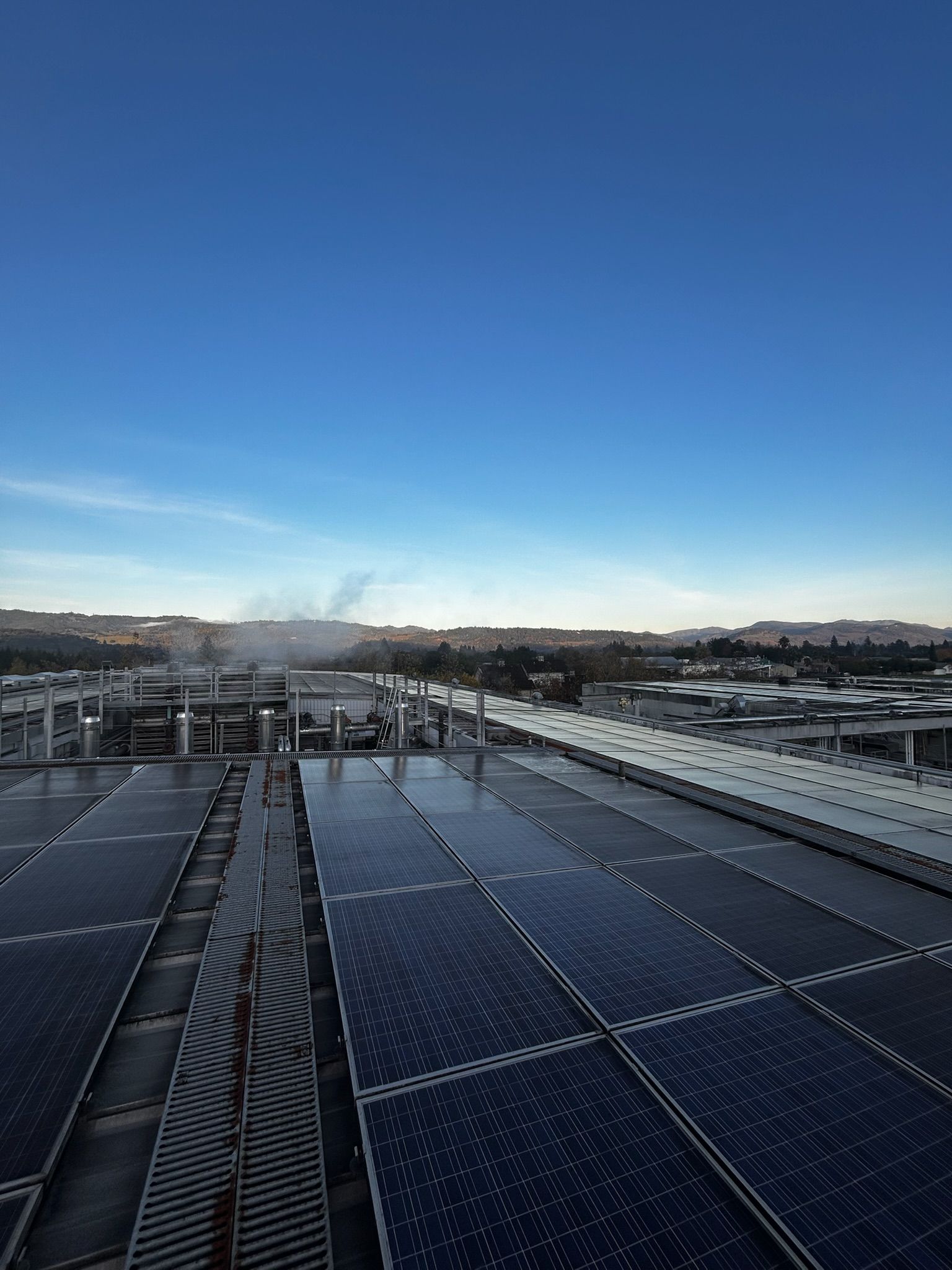 Rooftop with solar panels under a blue sky, with buildings and a hazy horizon in the distance.