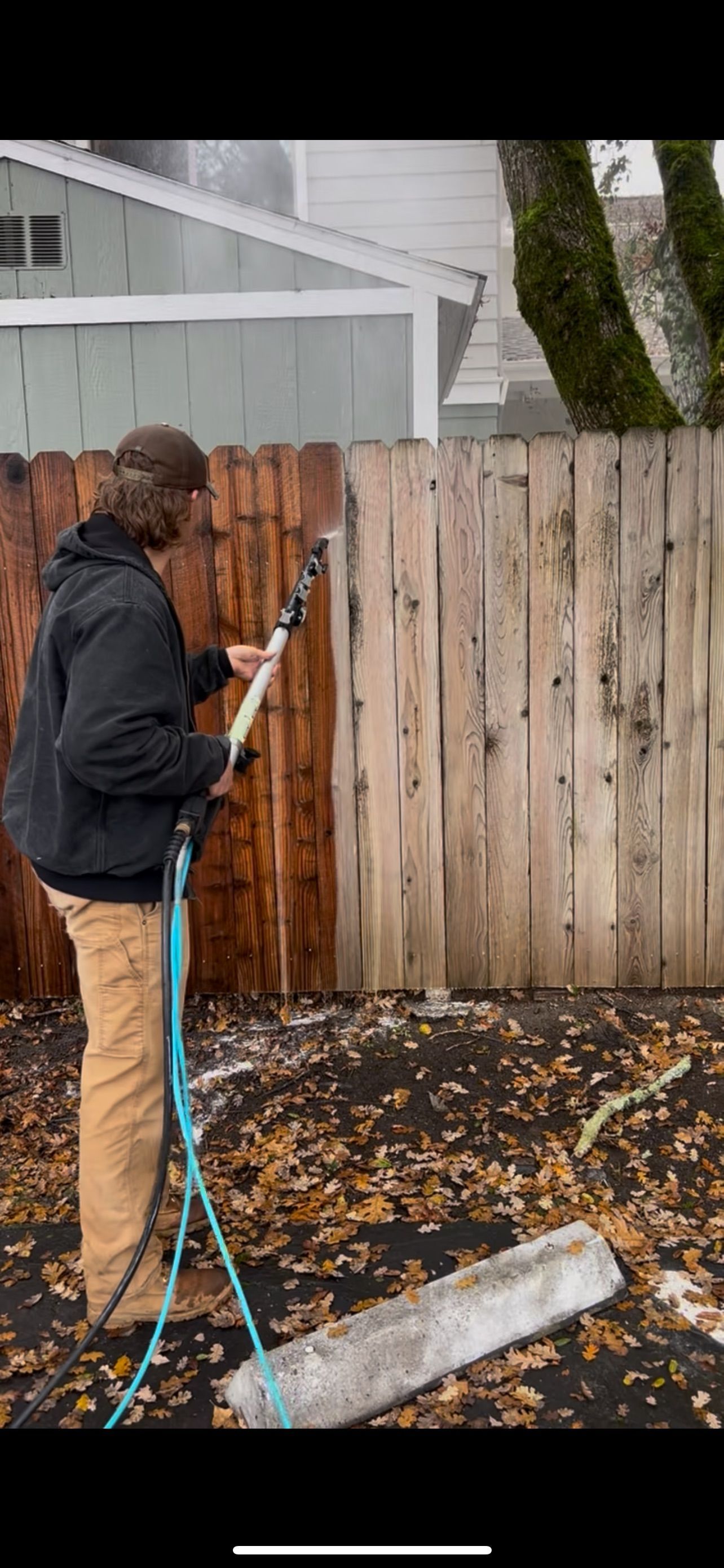 A person power washing a wooden fence, half clean, half dirty, in a yard with leaves.