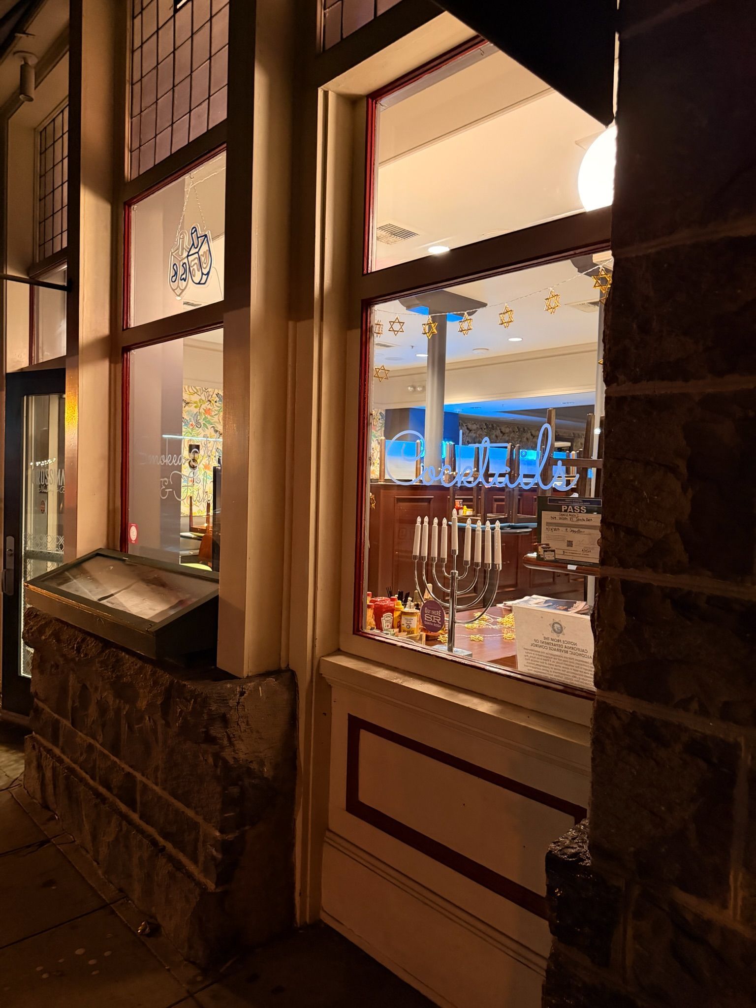 Exterior view of a brightly lit shop window at night, displaying candles and interior. Stone facade.