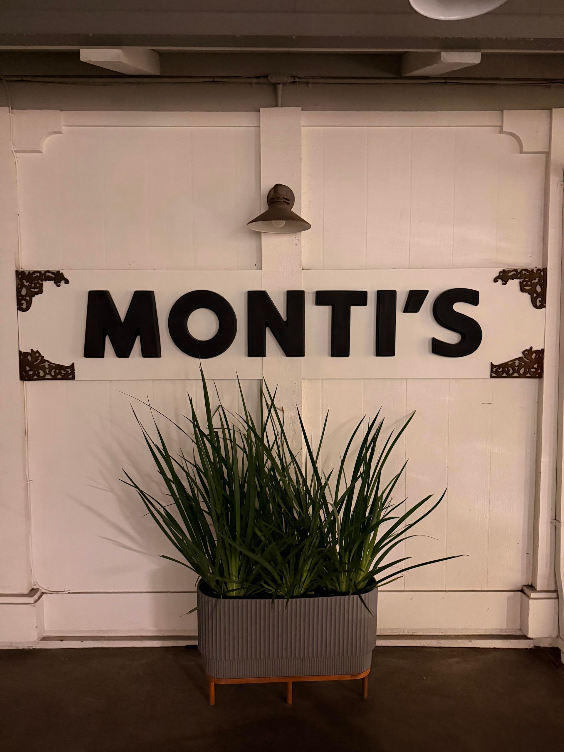Monti's restaurant sign on a white wall with a plant in a rectangular planter.