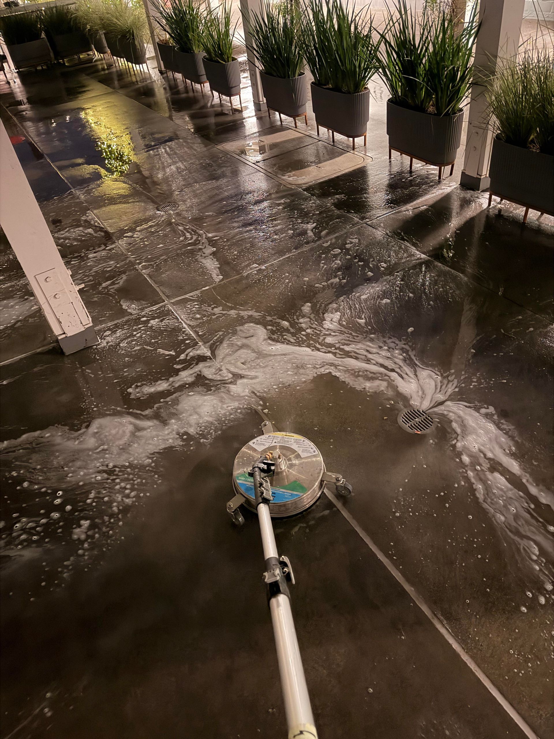 A person pressure washing a concrete patio, creating foamy water. Planter boxes line the background.