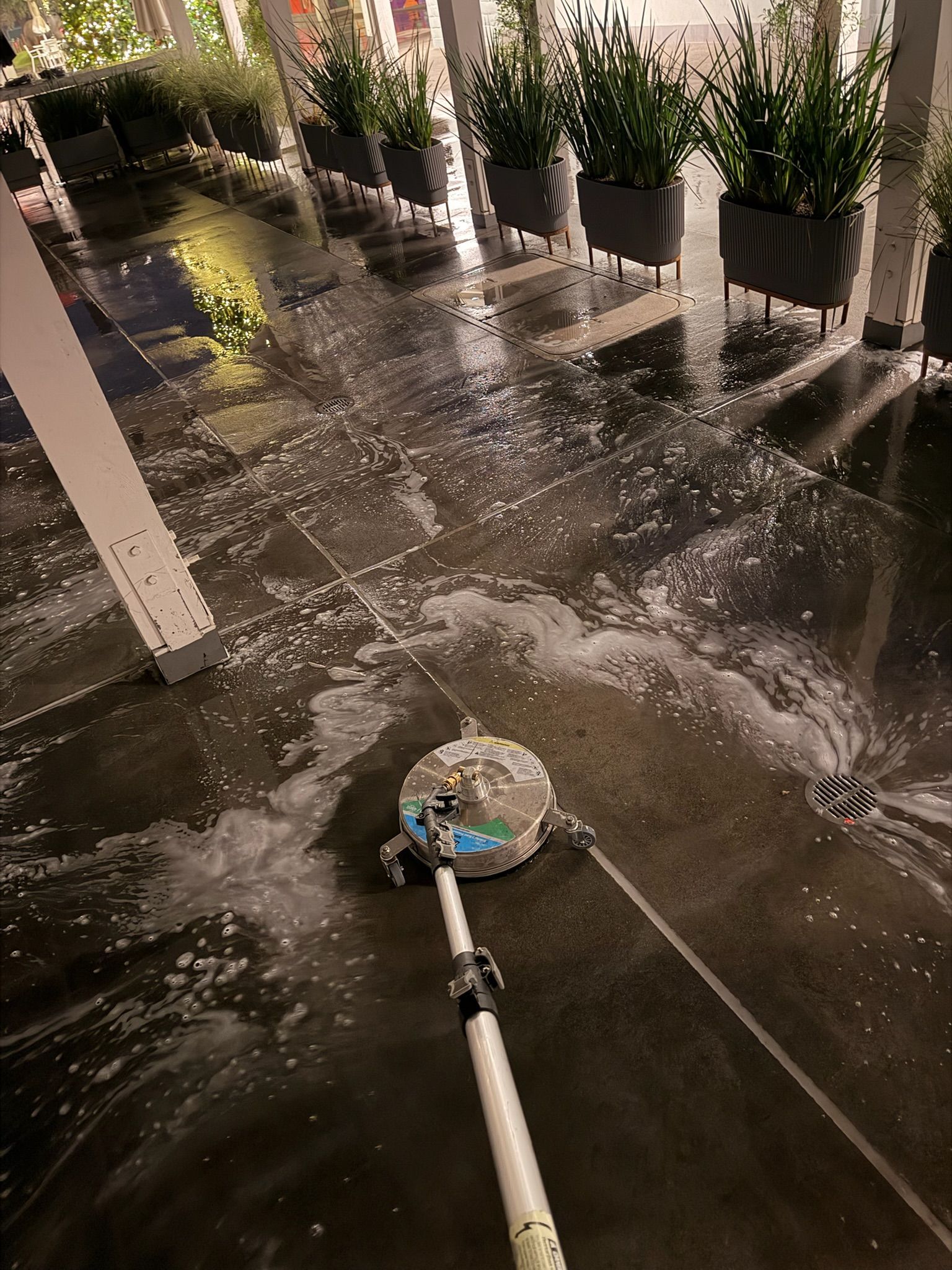 A person uses a power washer to clean a dark, wet outdoor patio with potted plants.