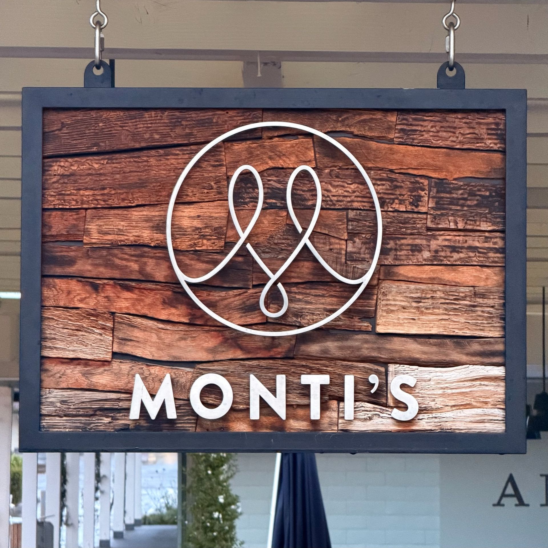 Sign for Monti's restaurant with white logo on wood background. Black frame, hanging from hooks.
