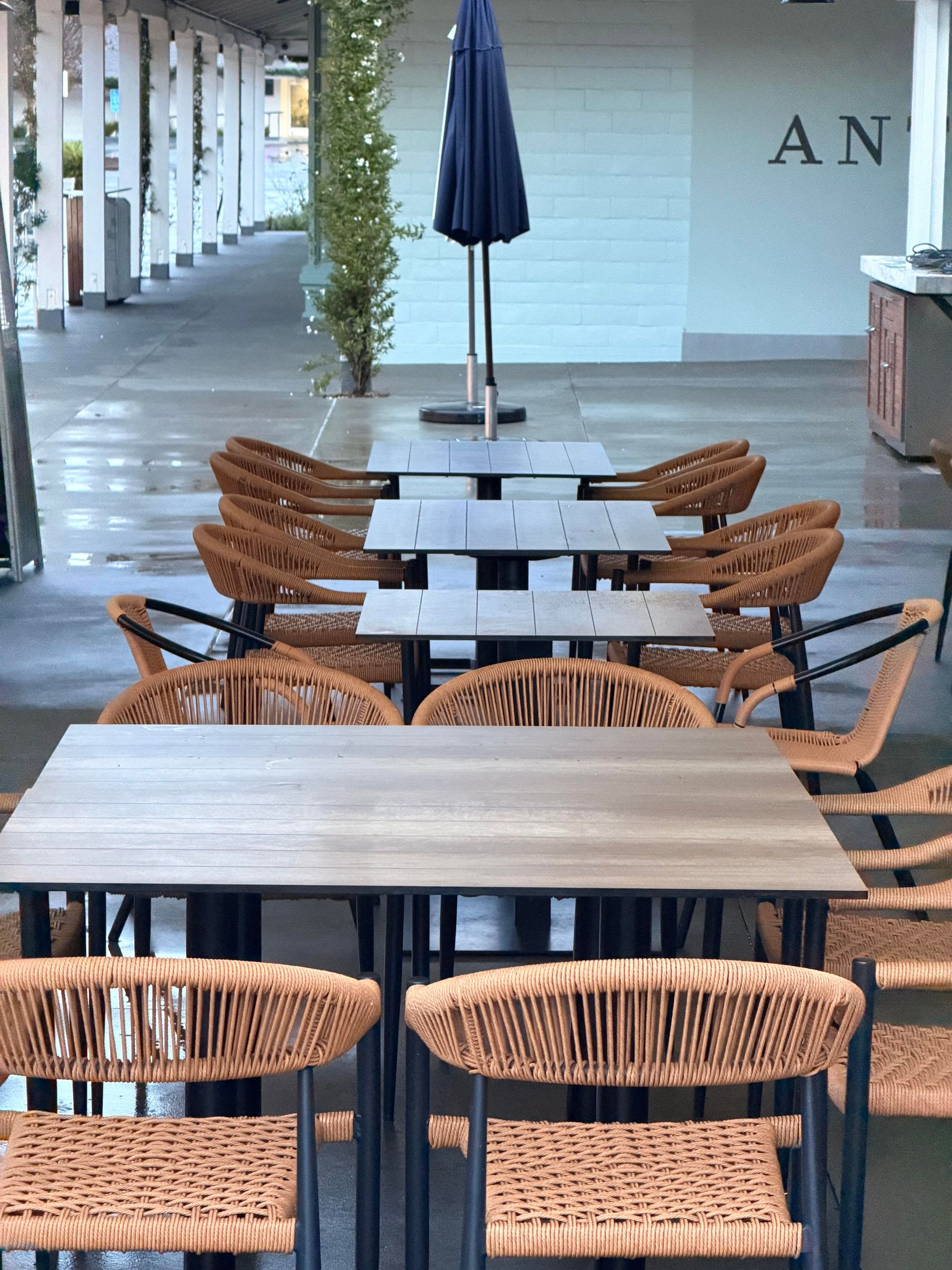 Outdoor dining area with woven chairs, square tables, and a closed umbrella.