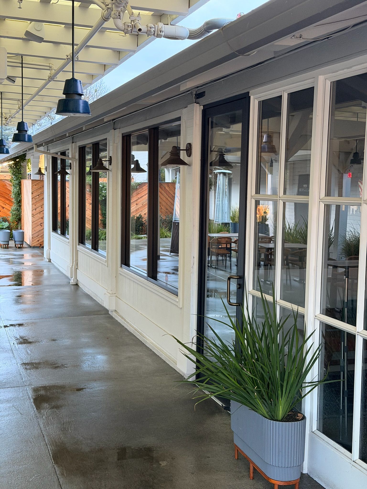 Exterior of a restaurant with white walls, black-framed windows, and hanging lights under a pergola.