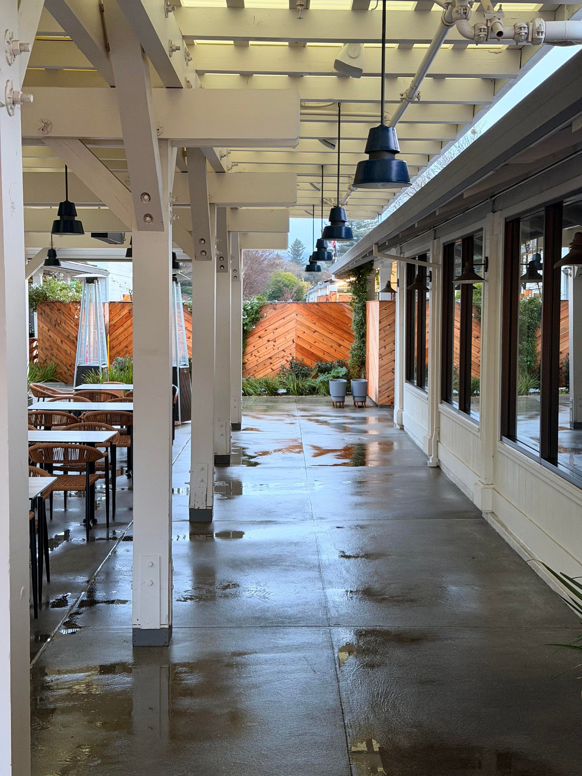 Covered outdoor restaurant patio with wet floor, tables, and blue pendant lights.