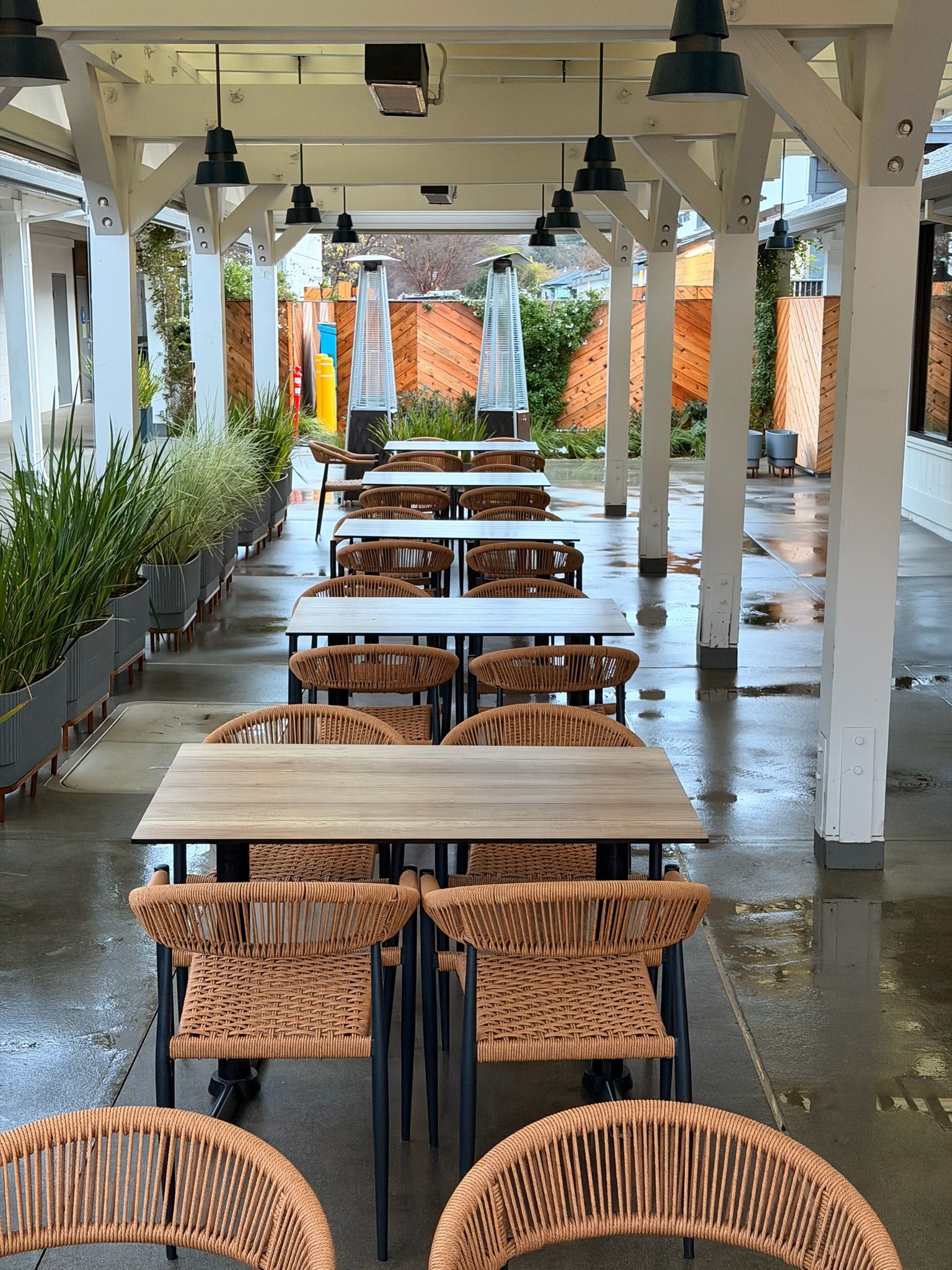 Outdoor restaurant seating under a white-framed awning. Tables and woven chairs are arranged in rows.