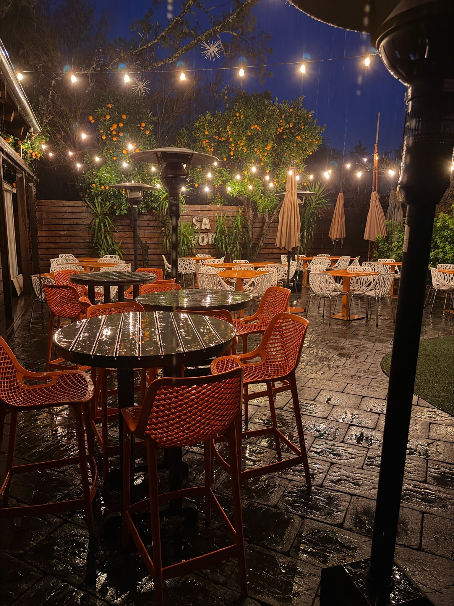 Outdoor restaurant patio at dusk with string lights and orange trees. Tables and chairs are wet from rain.