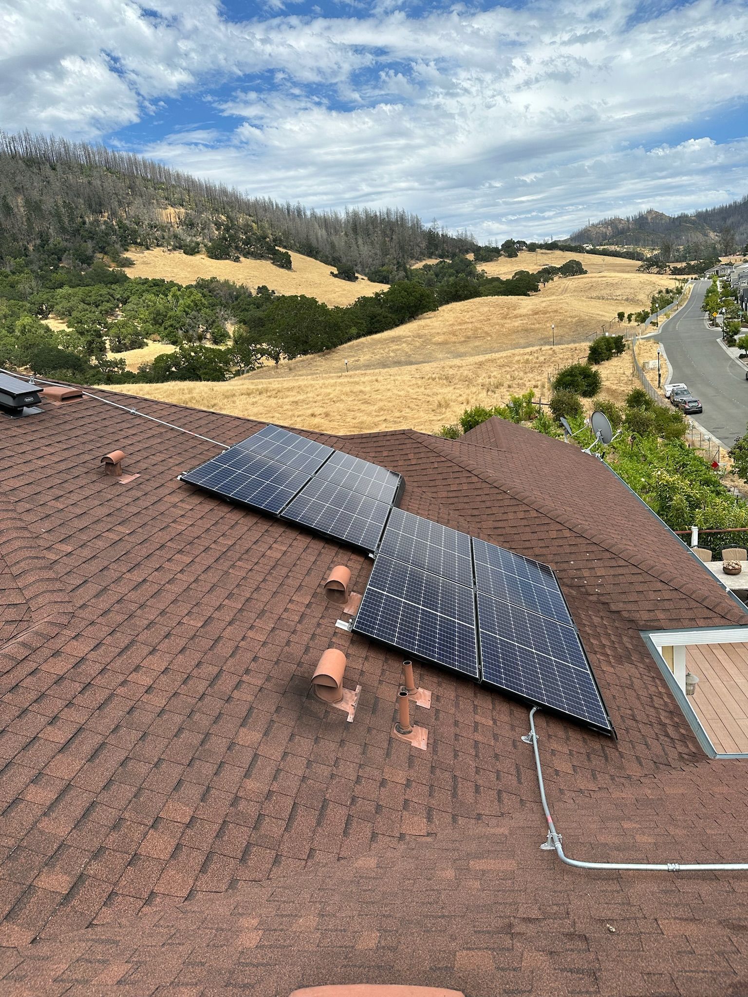 Solar panels on a brown-shingled roof overlooking a landscape of dry, grassy hills under a blue sky.