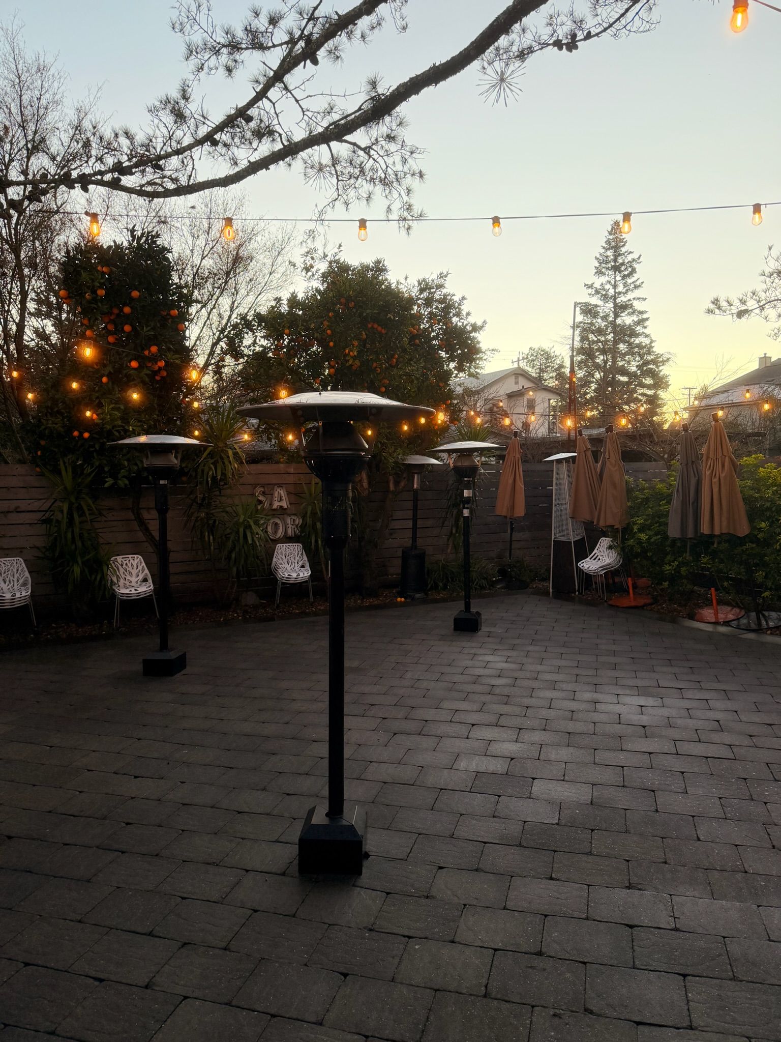 Patio with outdoor heaters, string lights, brick pavers, and trees at dusk.
