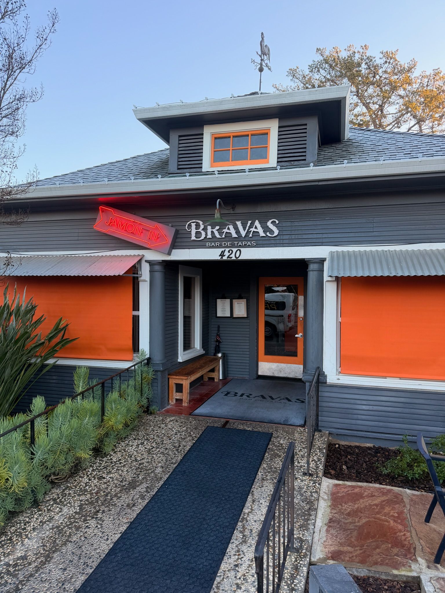 Exterior view of Bravas restaurant with grey siding, orange accents, and a neon sign.