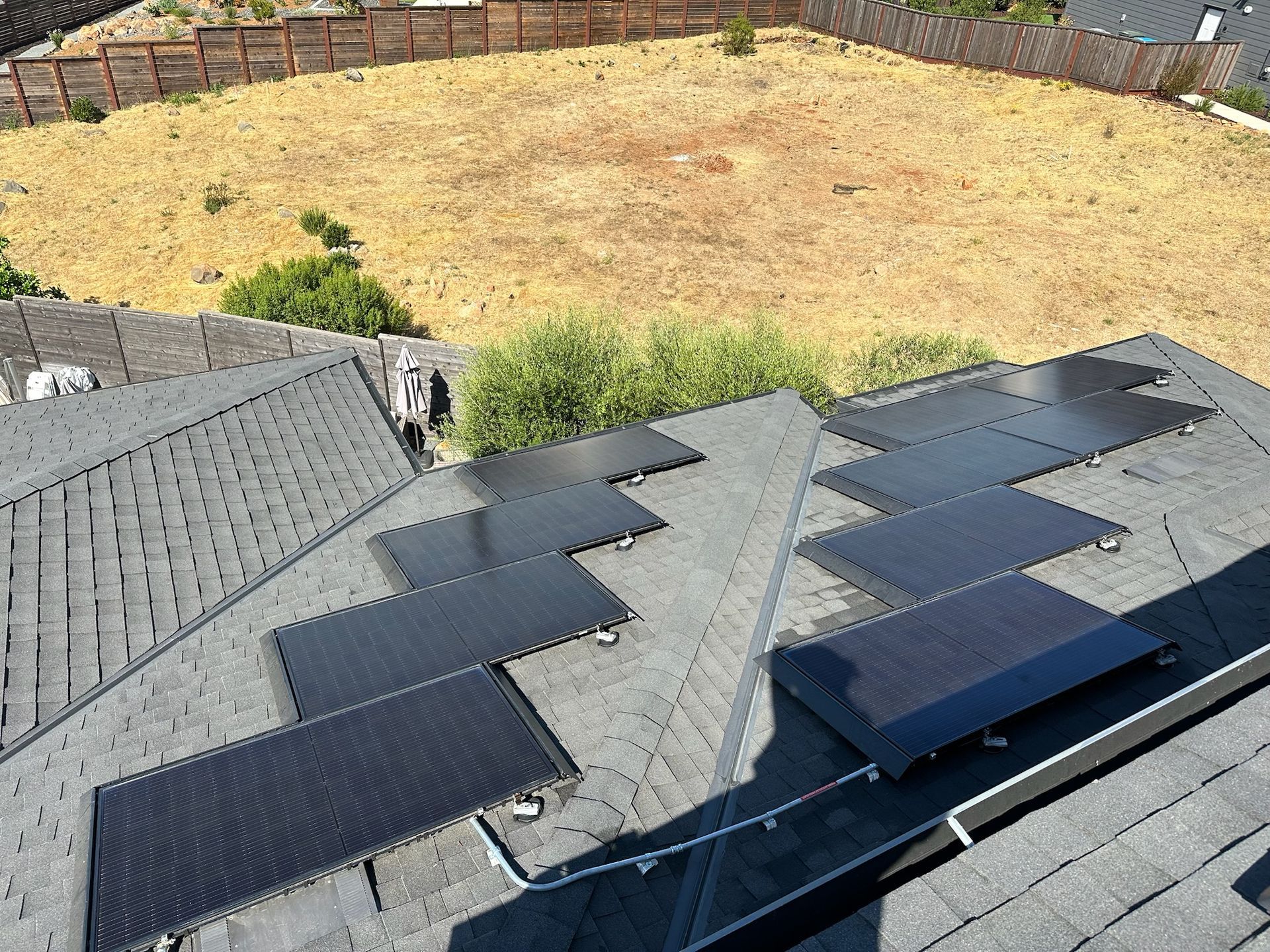 Solar panels installed on a dark grey shingle roof with a dry grass yard in the background.