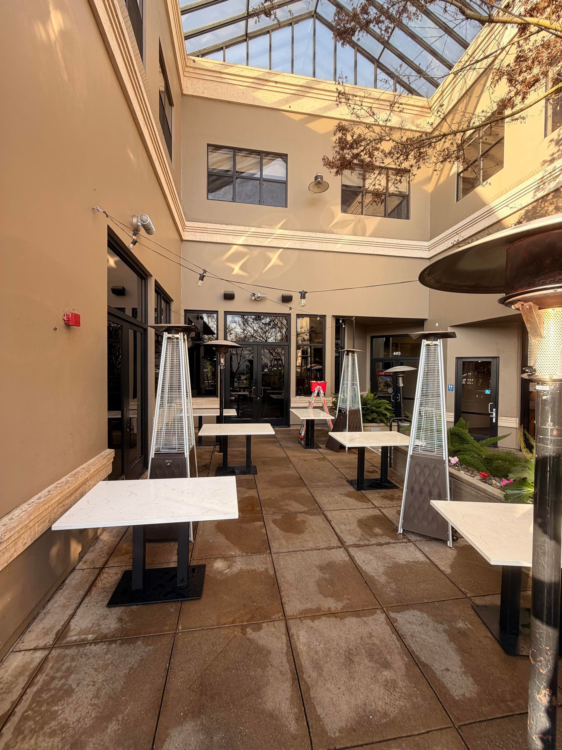 Outdoor patio with tables, heaters, and overhead glass roof. Restaurant entrance in background.