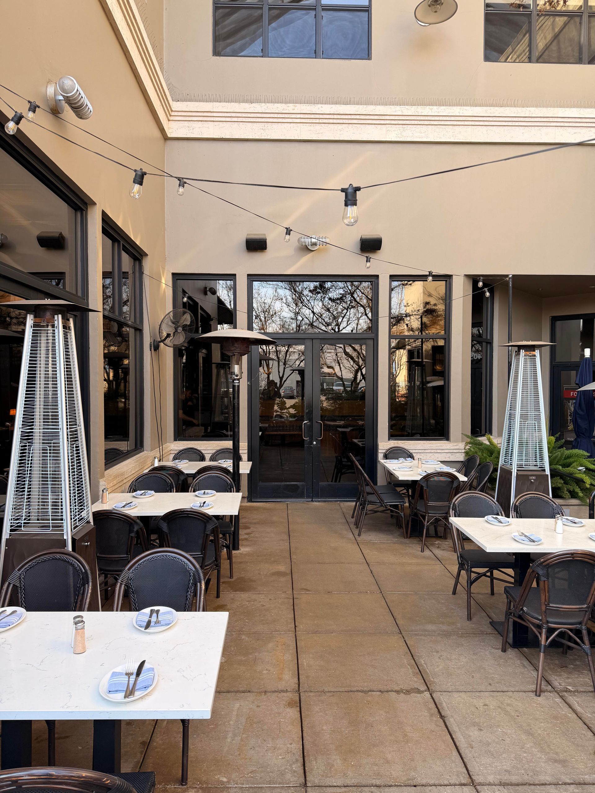 Outdoor restaurant patio with tables, chairs, and heaters. String lights overhead, dark glass doors in the background.