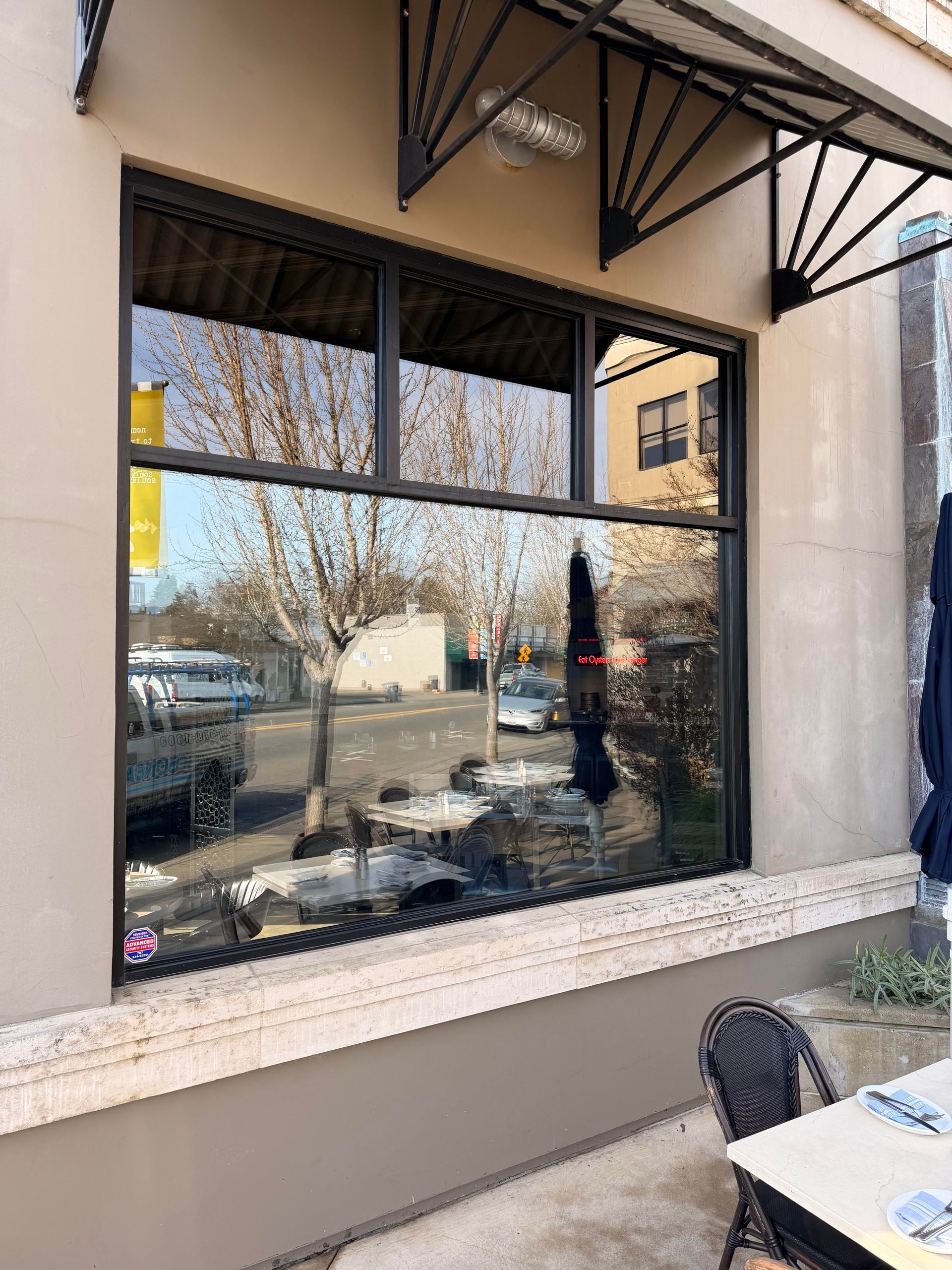 Large window of a building reflects the street.  A black awning hangs above, and there is a table and chair in front.