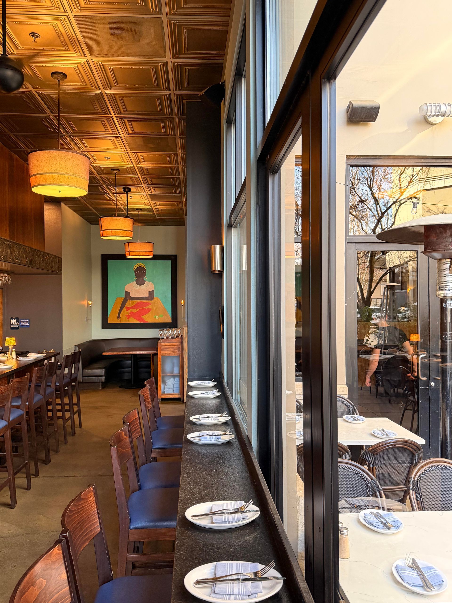 Restaurant interior with bar seating, a framed painting, and window tables overlooking an outdoor patio.