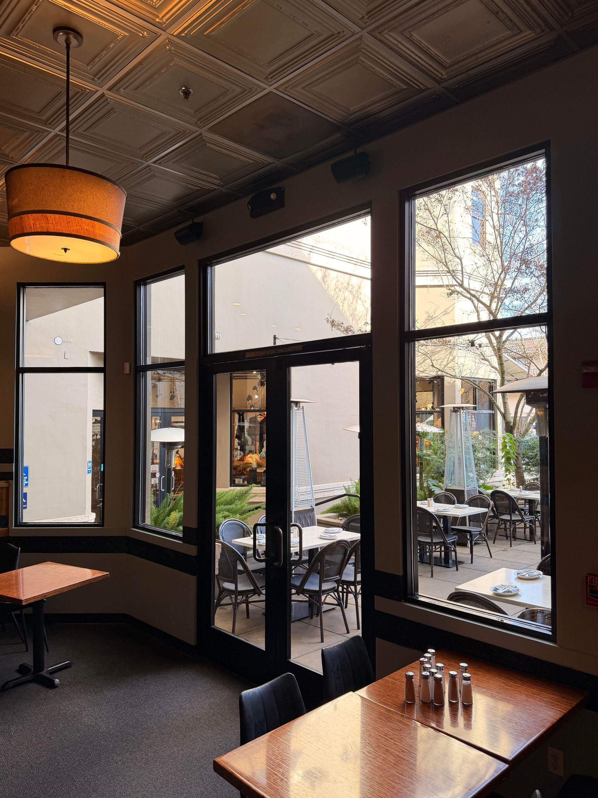 Restaurant interior with windows overlooking a patio with tables and chairs. Ceiling light and tables are visible.
