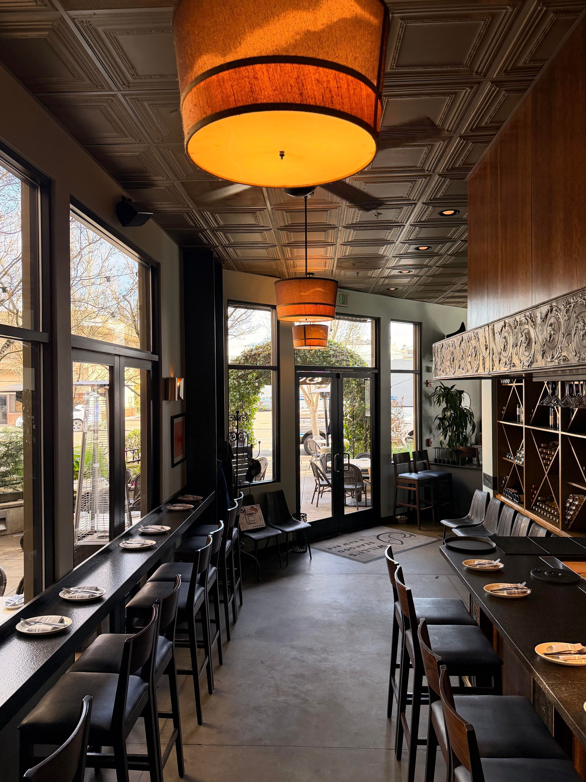 Restaurant interior with large windows, high chairs at a counter, and hanging orange lamps.