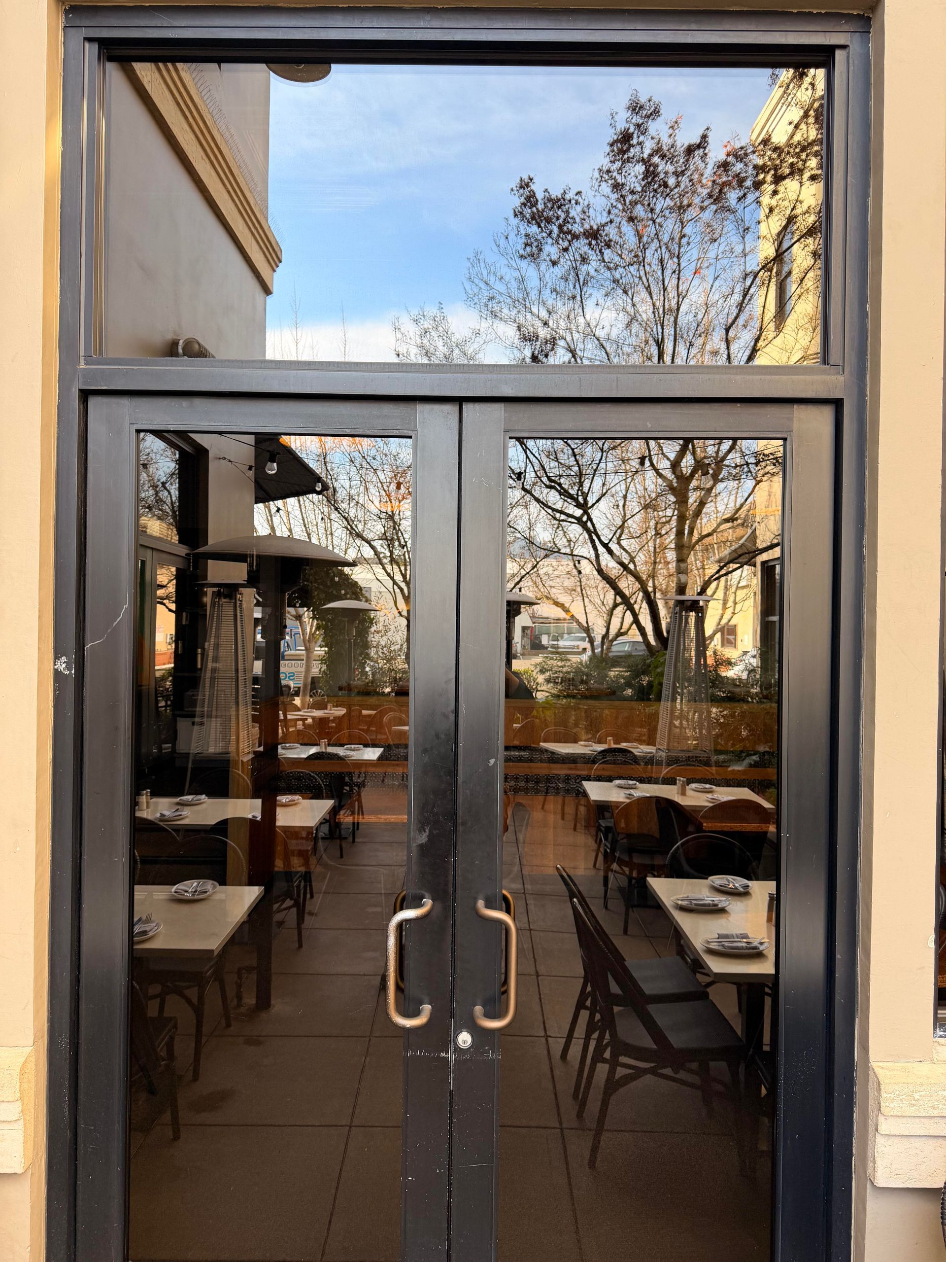 Exterior view of a restaurant. Glass doors reflect outdoor seating, trees, and blue sky.