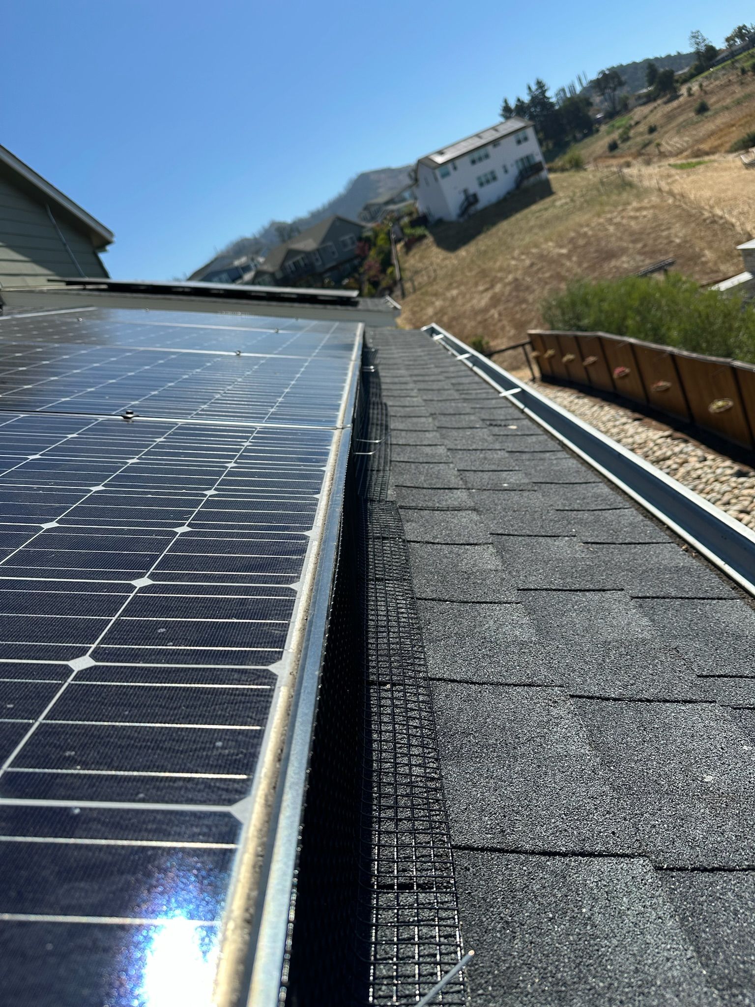 Solar panel on dark roof next to shingles, with hillside and building in background under blue sky.