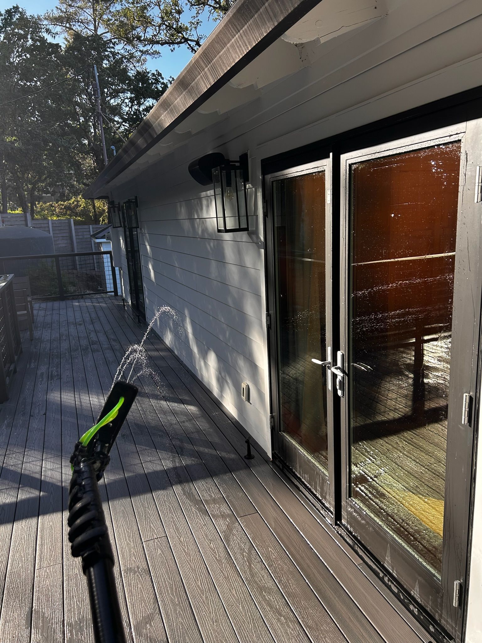 A person using a pressure washer on a gray deck next to a building with black-framed glass doors.