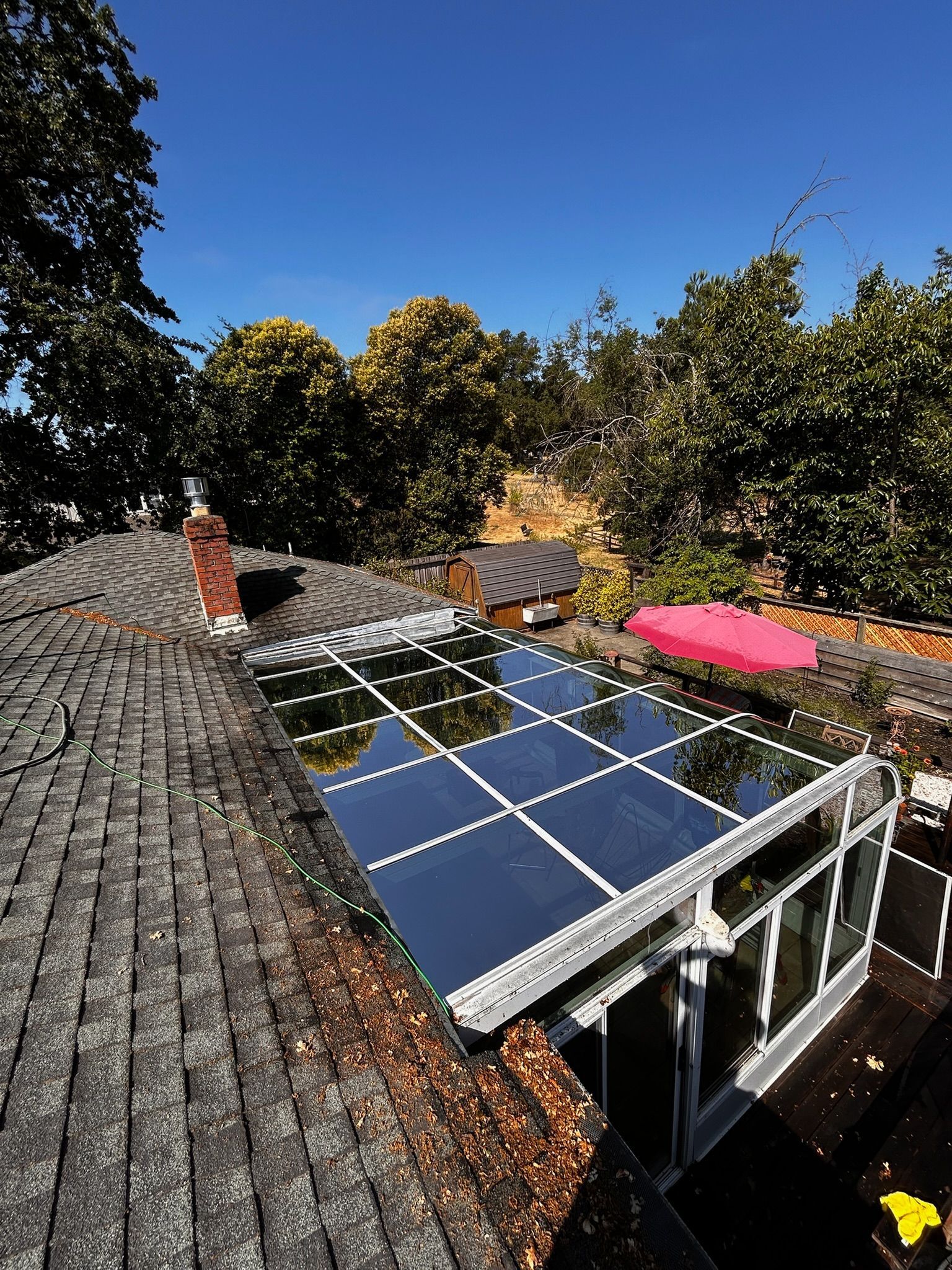 Rooftop view of solar panels on a glass enclosure, surrounded by trees under a clear blue sky.