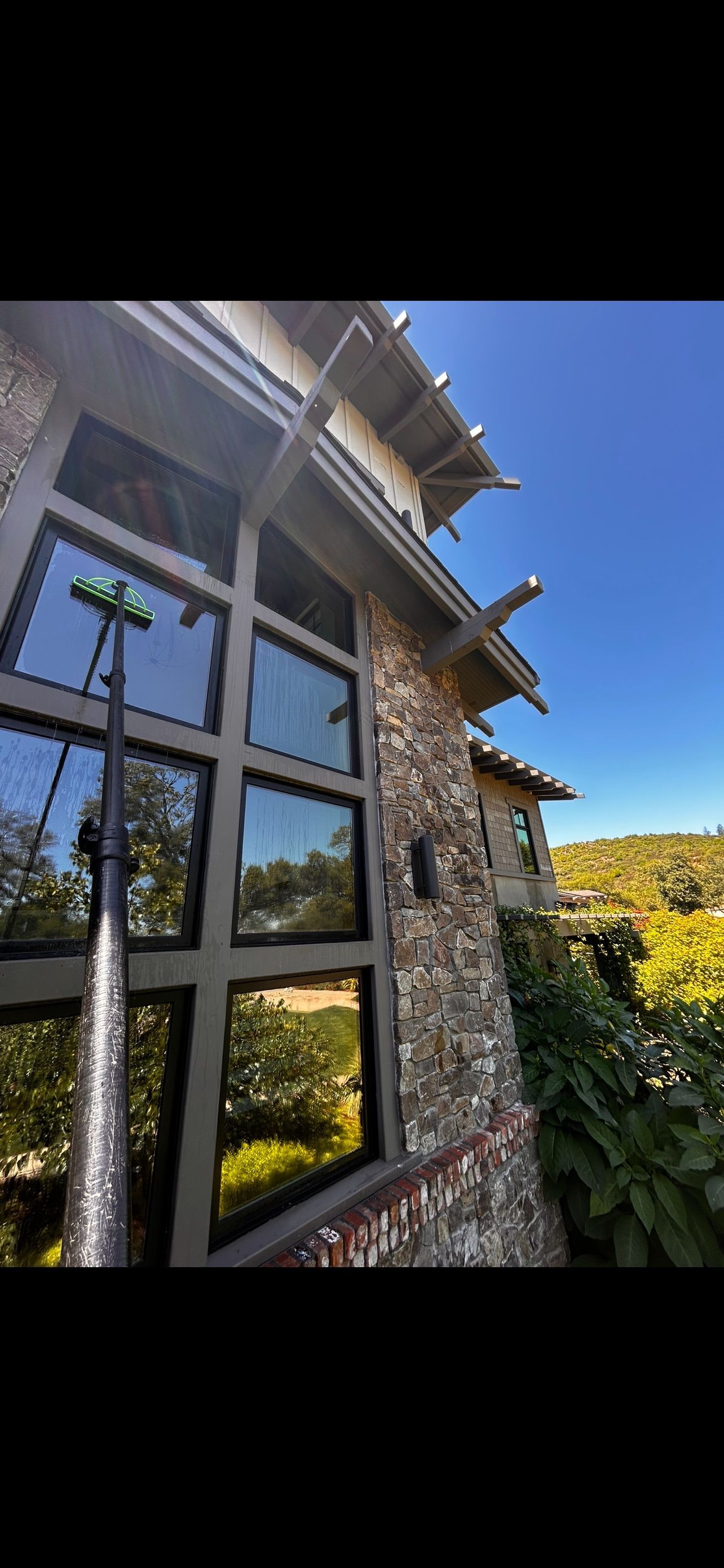 A person washing windows on a stone building with a water-fed pole. Blue sky and greenery in the reflection.