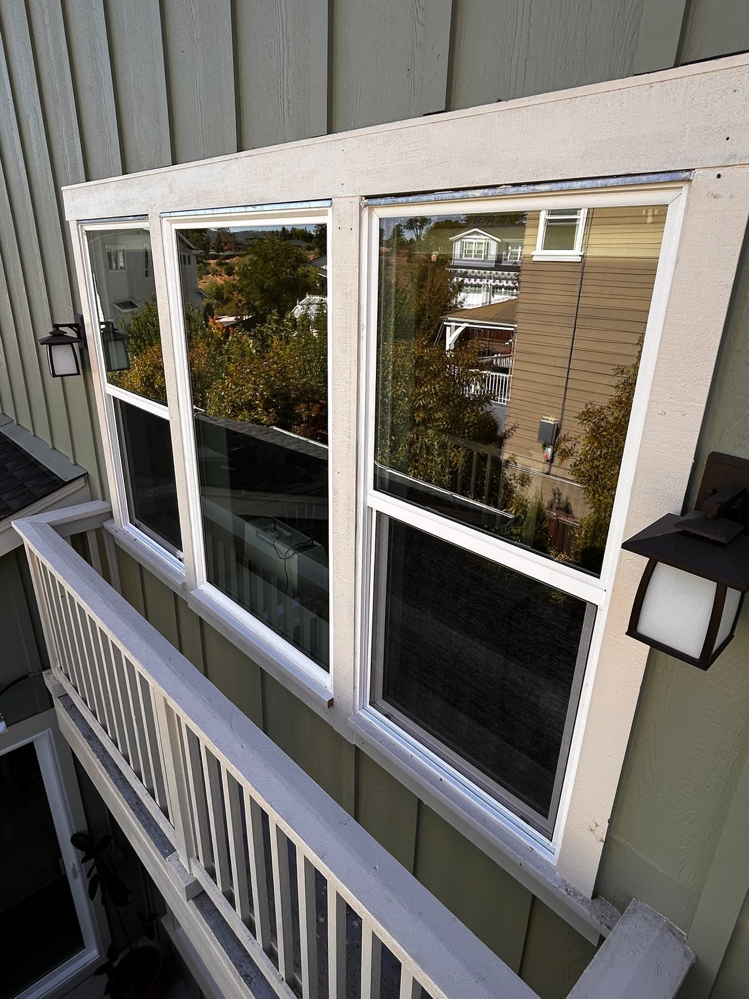 Three-panel window on green building with white trim, reflecting trees and buildings. Railing below, exterior lights.