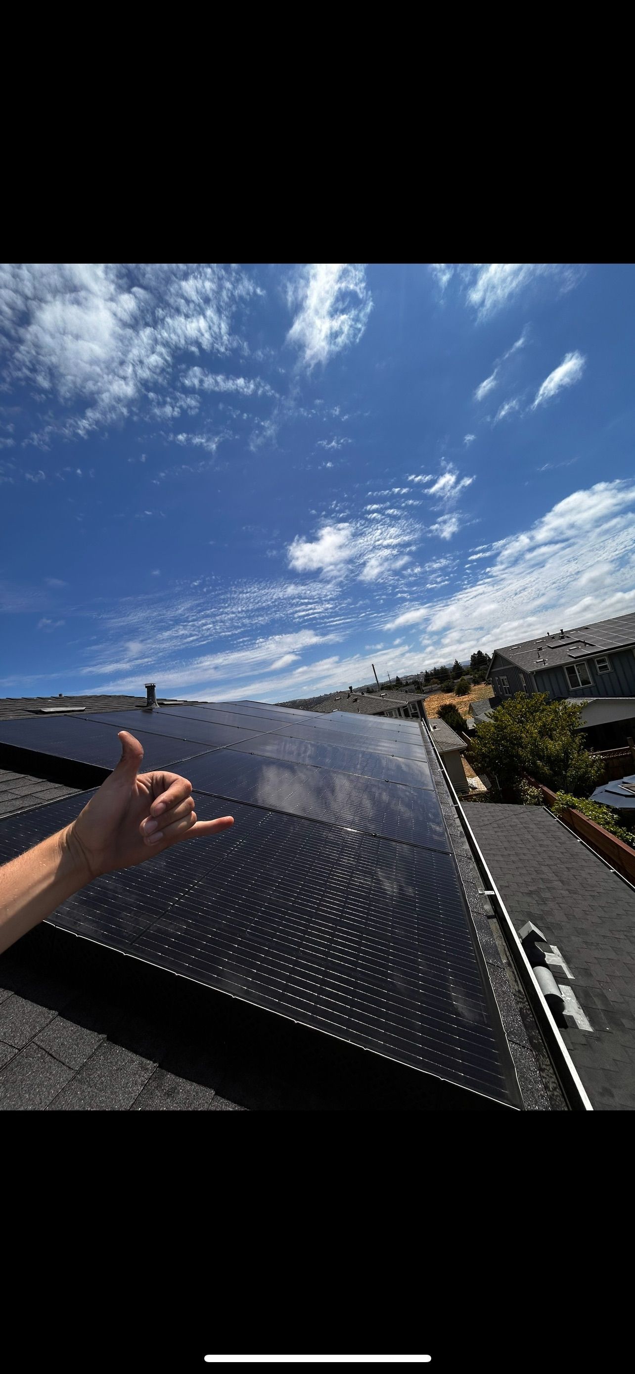 Person giving a shaka sign in front of solar panels on a rooftop with a bright blue sky.