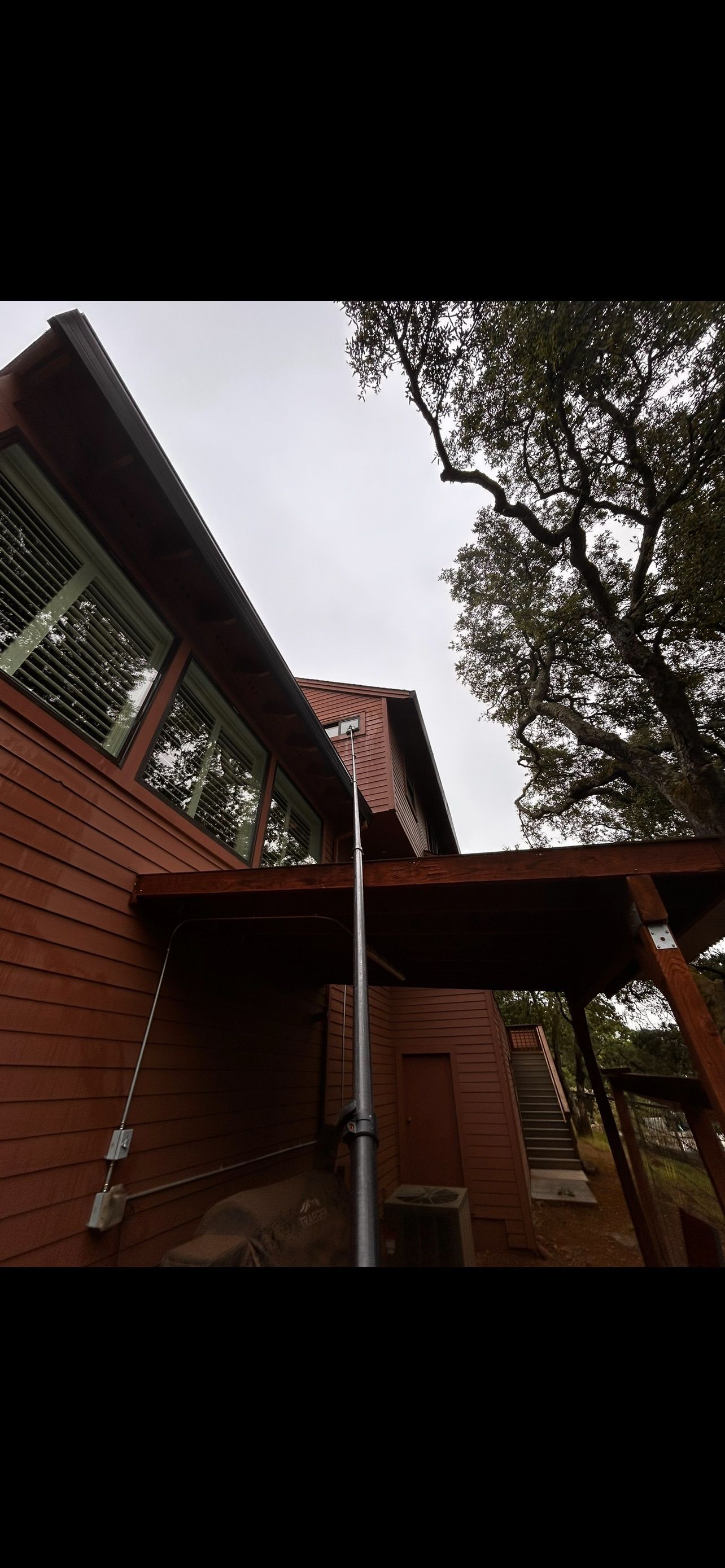 Exterior view of a red wooden house with large windows, tall tree and a metal pole in the center. Overcast sky.
