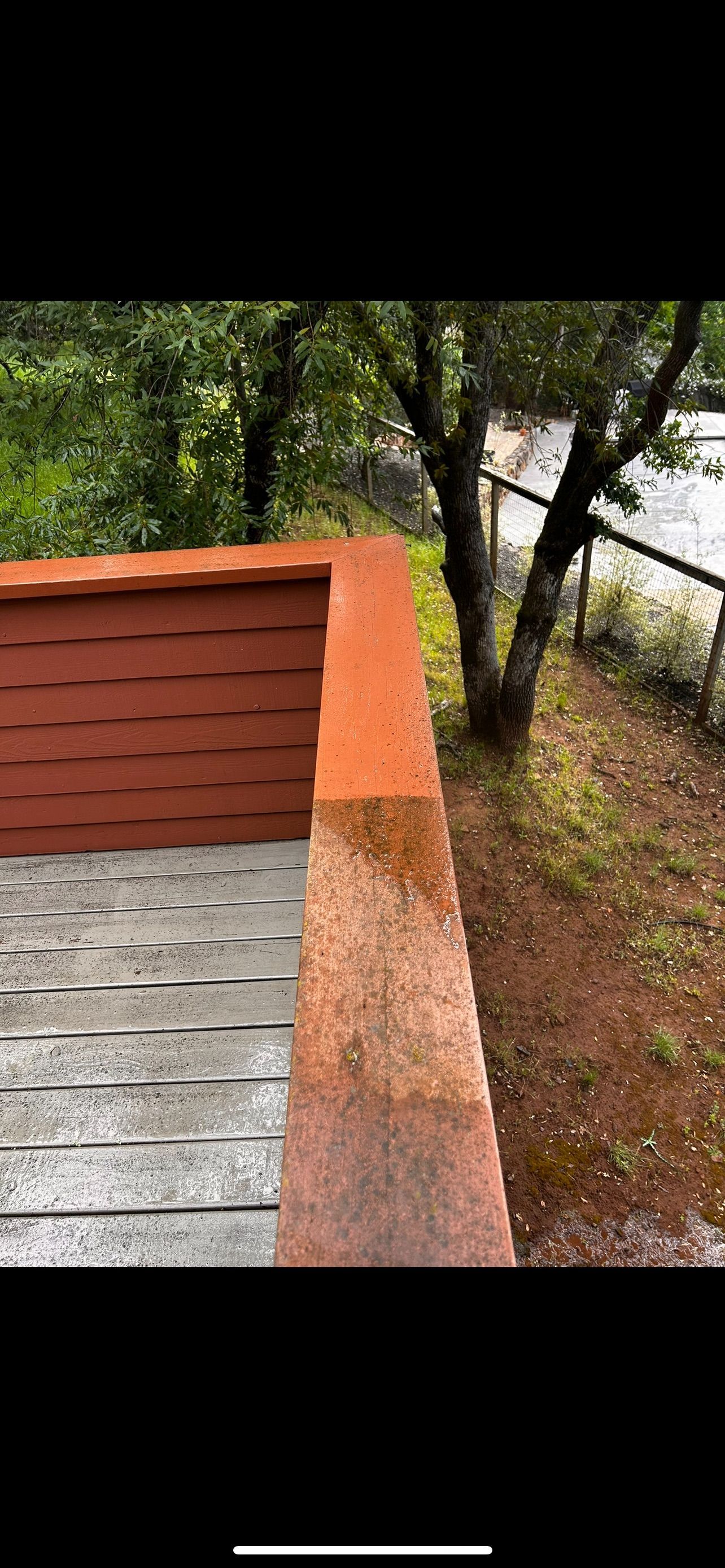 View from a deck with reddish-brown railing and siding, overlooking trees and a grey fence, rainy day.