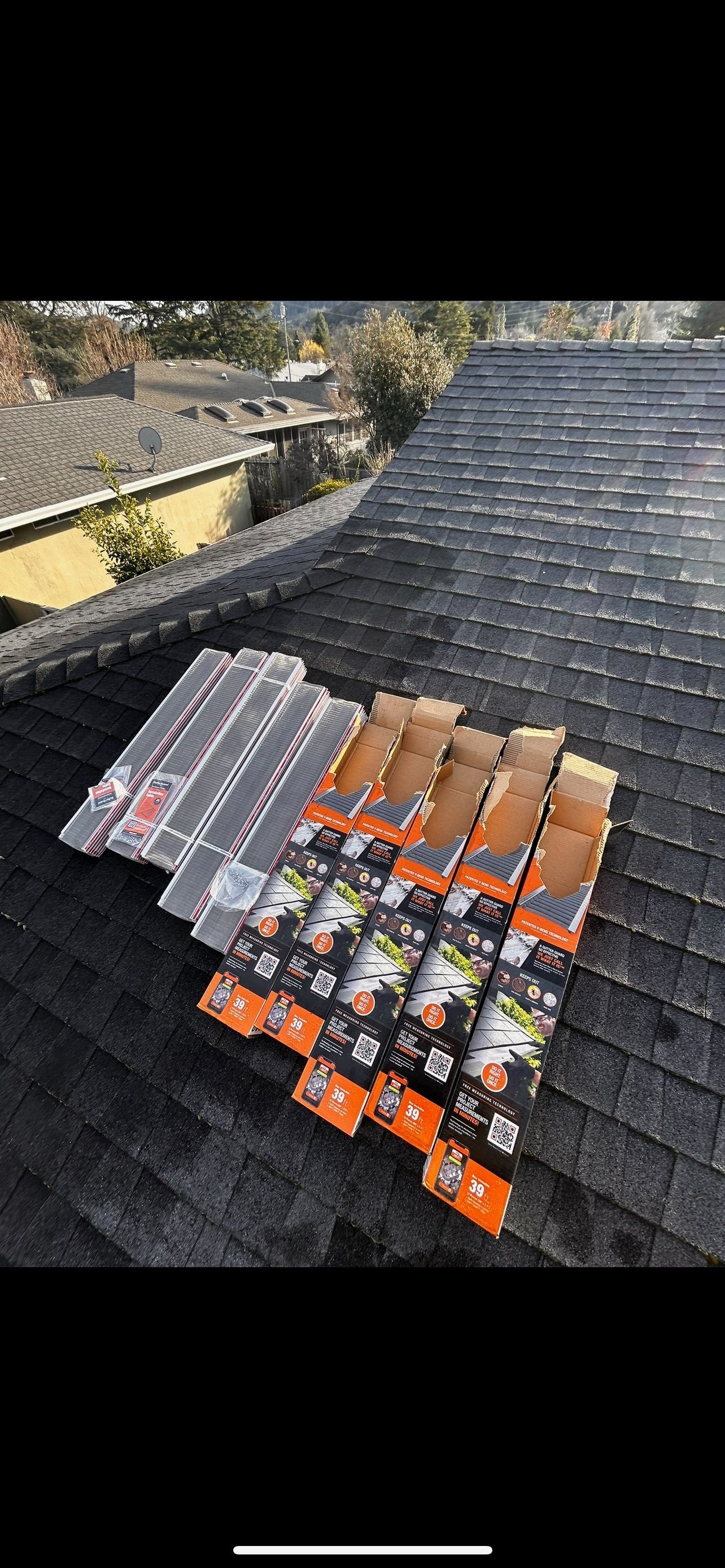 Various boxes of roofing materials arranged on a dark shingled roof, trees and other buildings visible in background.