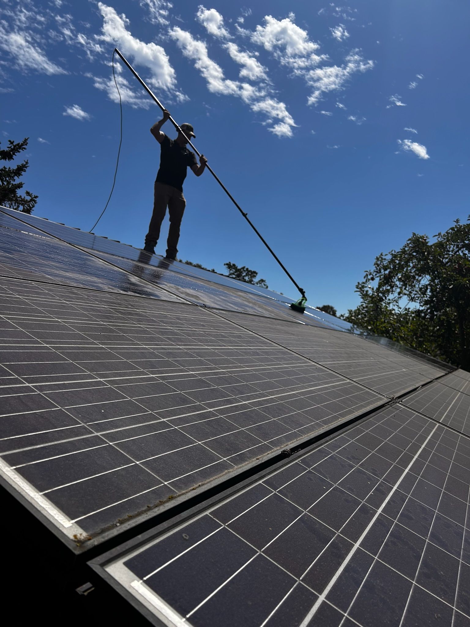 Person cleaning solar panels on a roof using an extended brush under a bright blue sky.