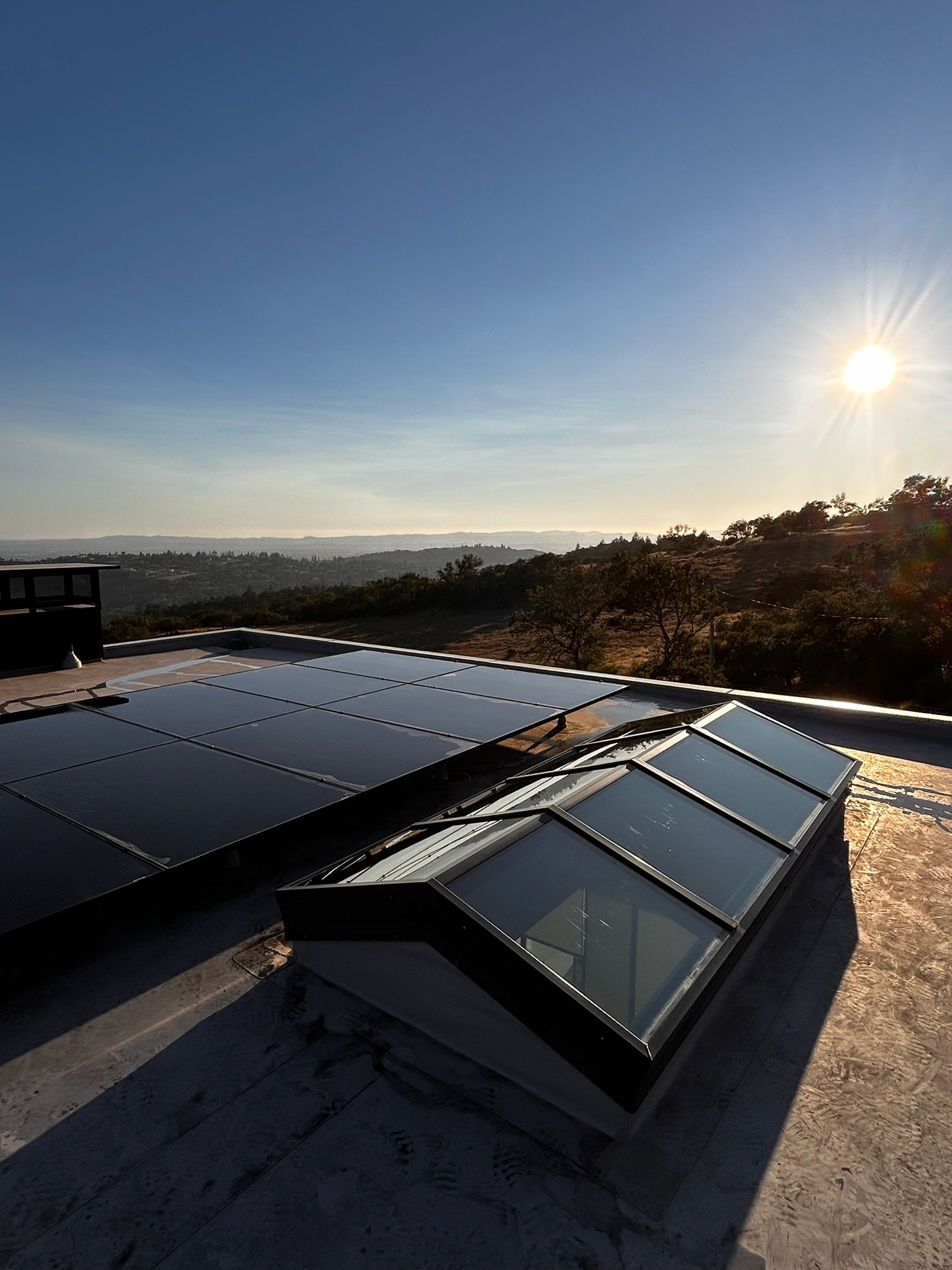 Solar panels and skylight on a flat roof, overlooking a hilly landscape under a bright sun.