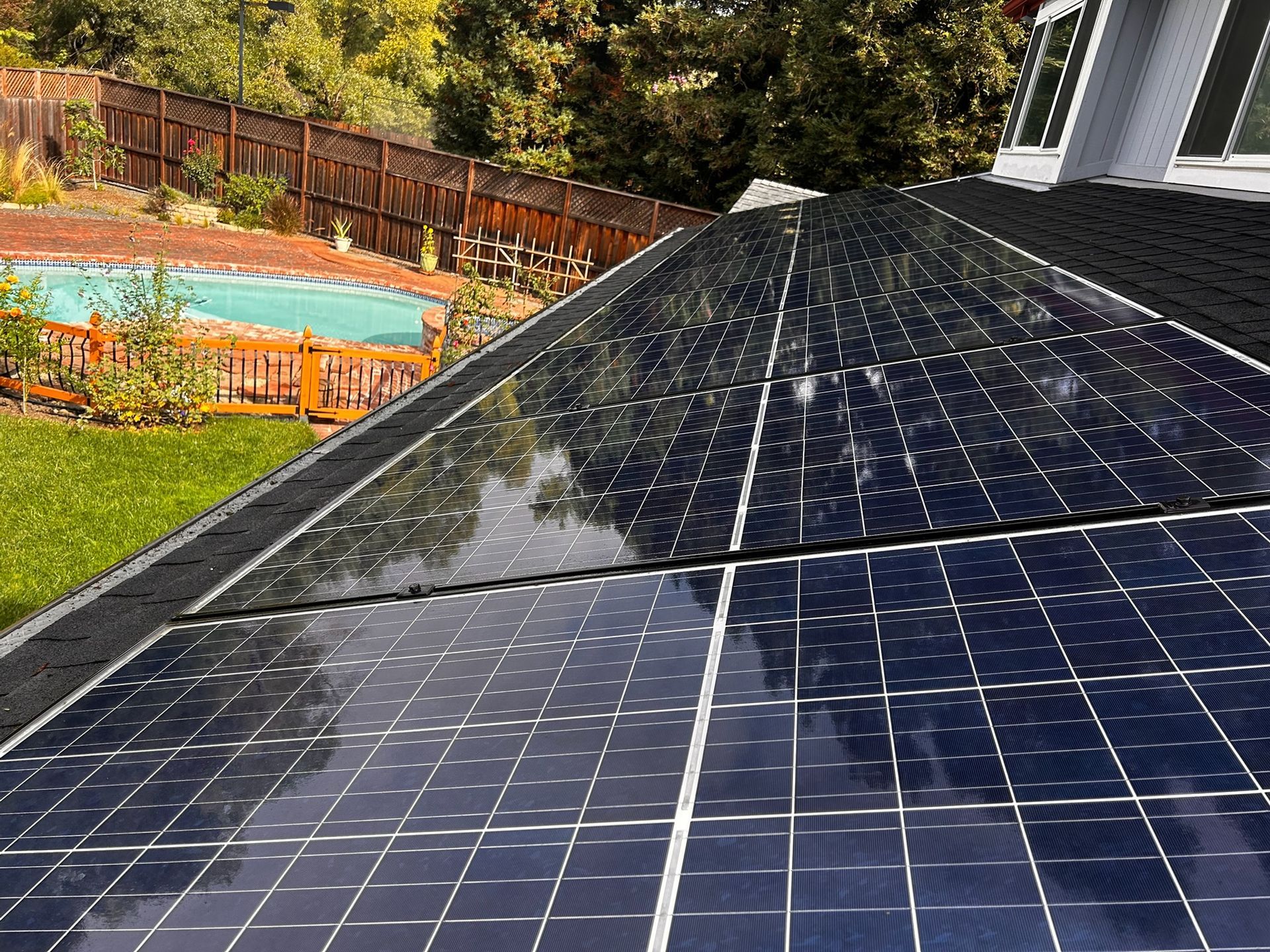 Solar panels on a roof reflecting the sky, with a backyard pool and fence in the background.