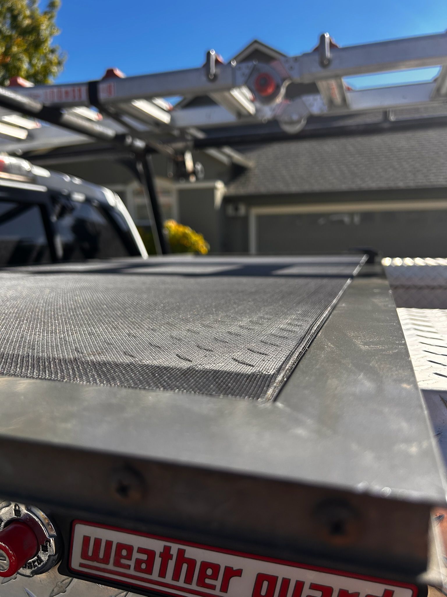 Close-up of a Weather Guard truck bed cover with a ladder rack in the background. Blue sky and a house are visible.