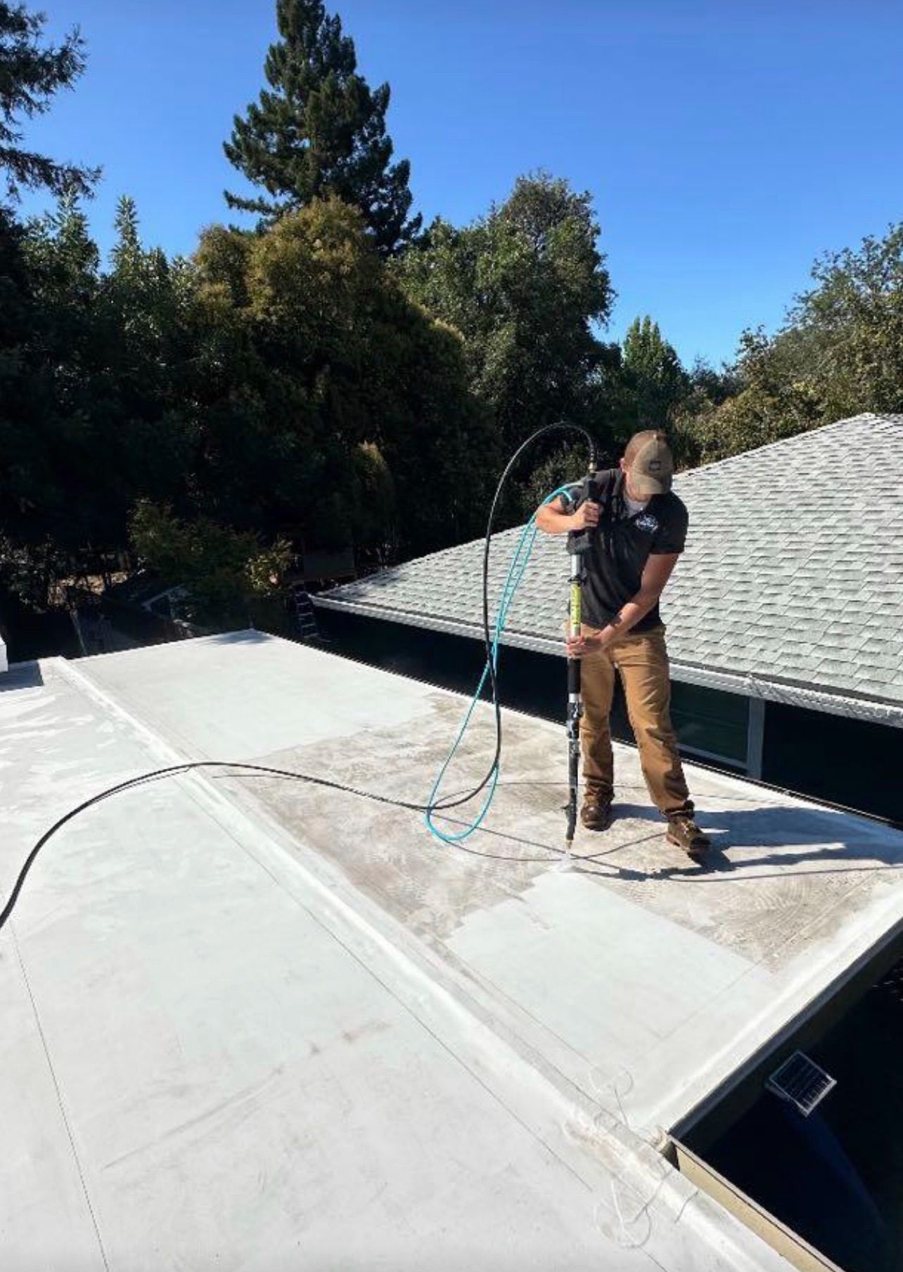 A person on a rooftop uses a hose, possibly cleaning, with trees and a blue sky in the background.