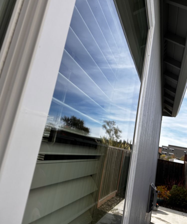 Angled window reflecting a blue sky with clouds, a fence, and parts of a building.
