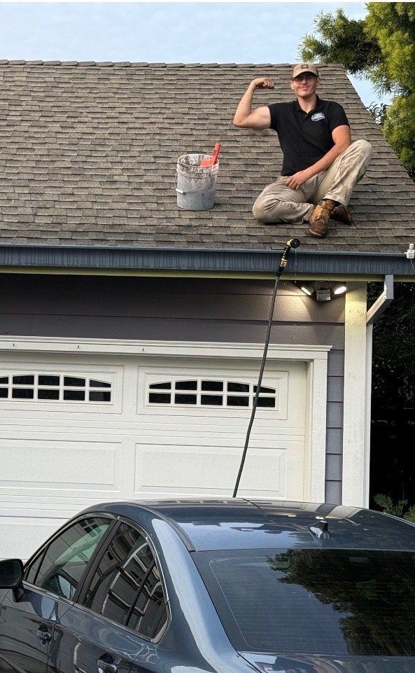 Man flexing arm on roof, cleaning gutters with long-handled tool. Bucket and car in view.