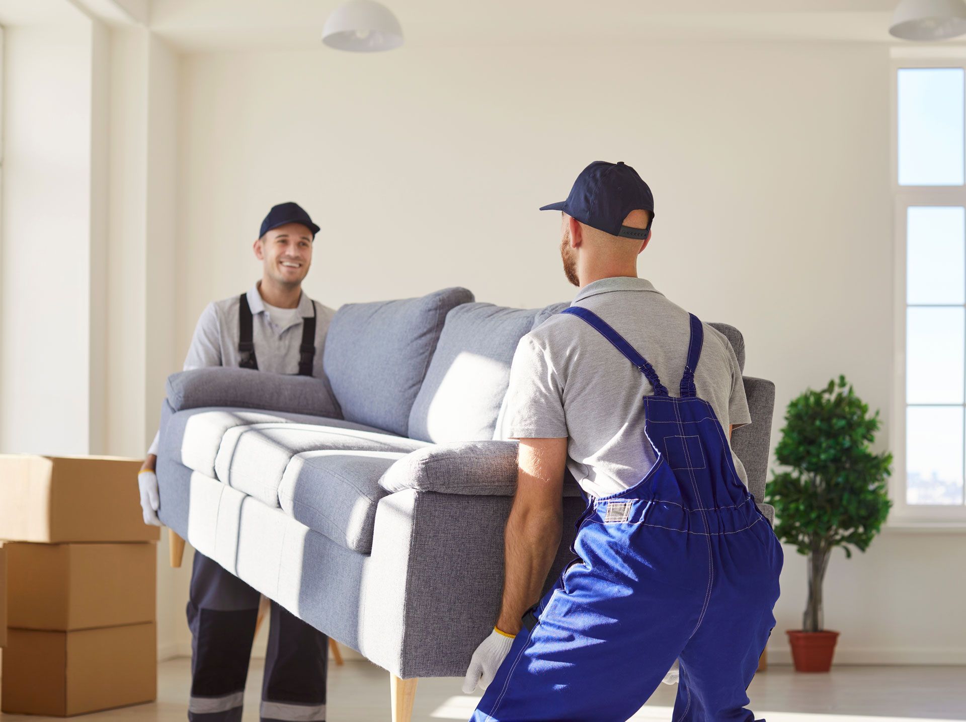 Two movers in blue coveralls carrying a gray couch in a bright room.