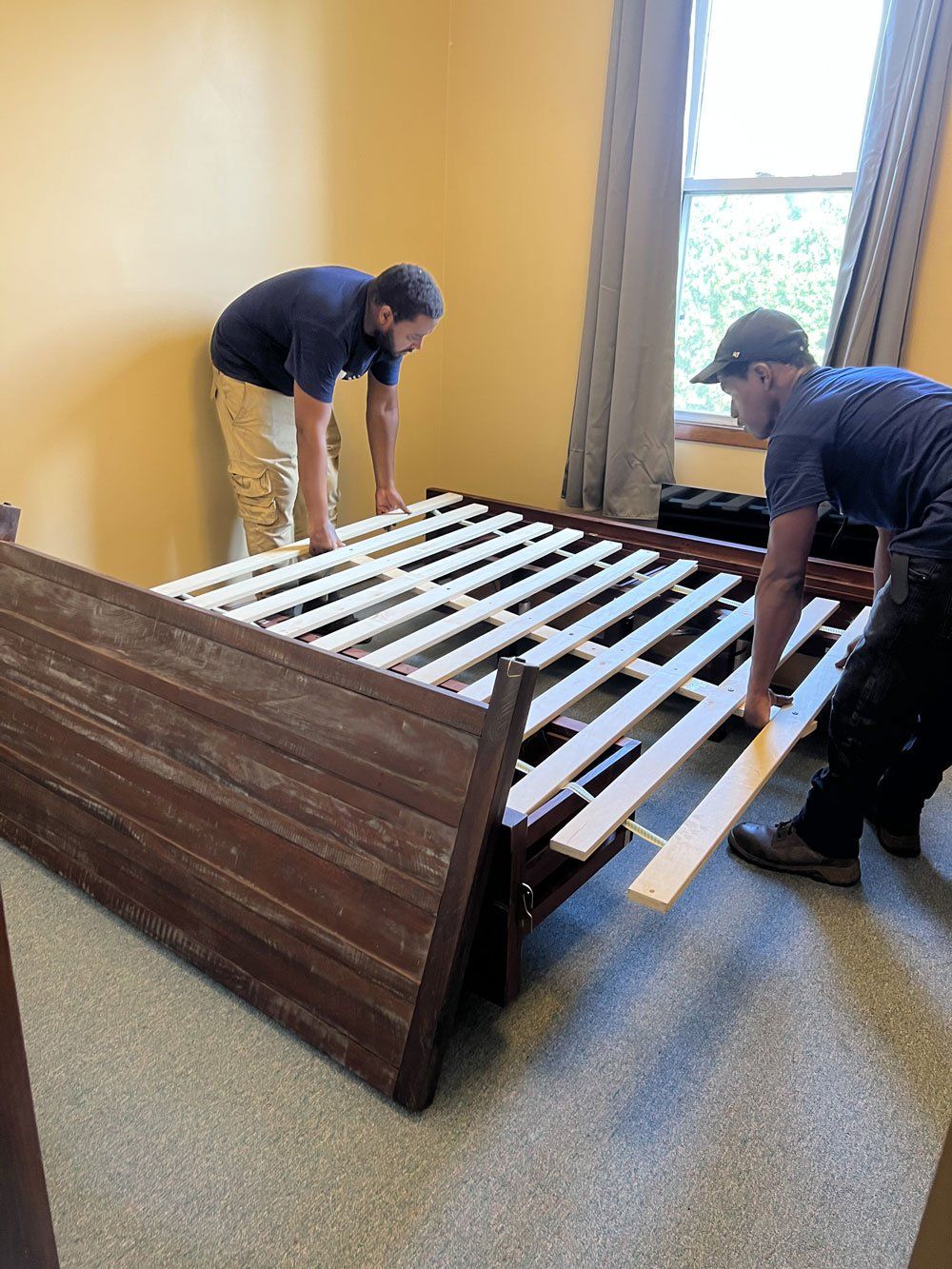 Two men are working on a wooden bed frame in a bedroom.