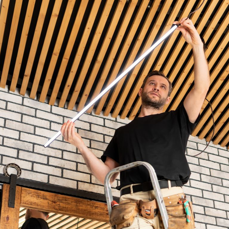 Man on a ladder installing a long, white LED light fixture onto a wooden ceiling.