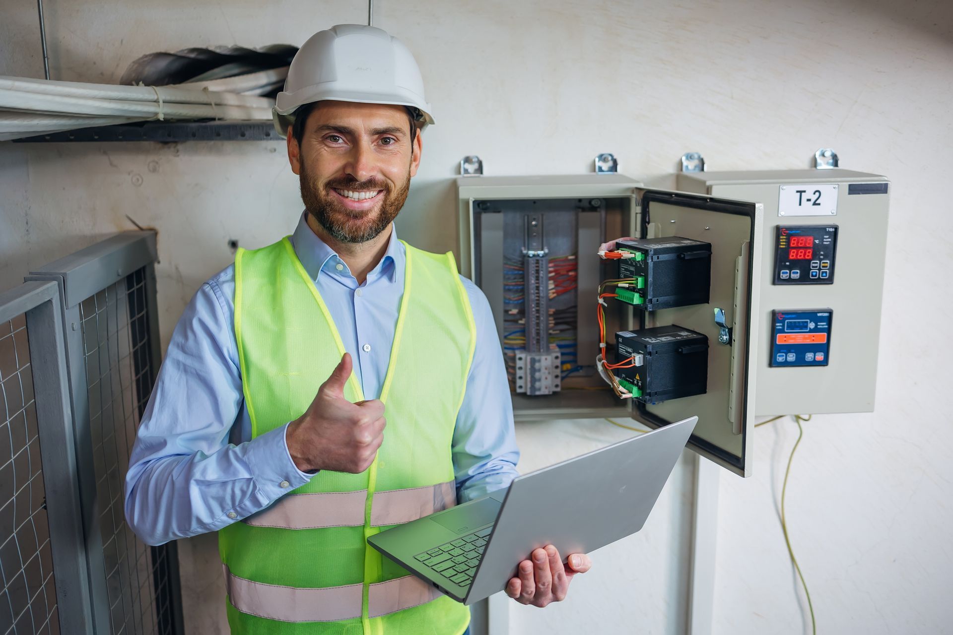 Smiling electrician in safety vest and hard hat giving thumbs up, holding laptop near electrical panel.