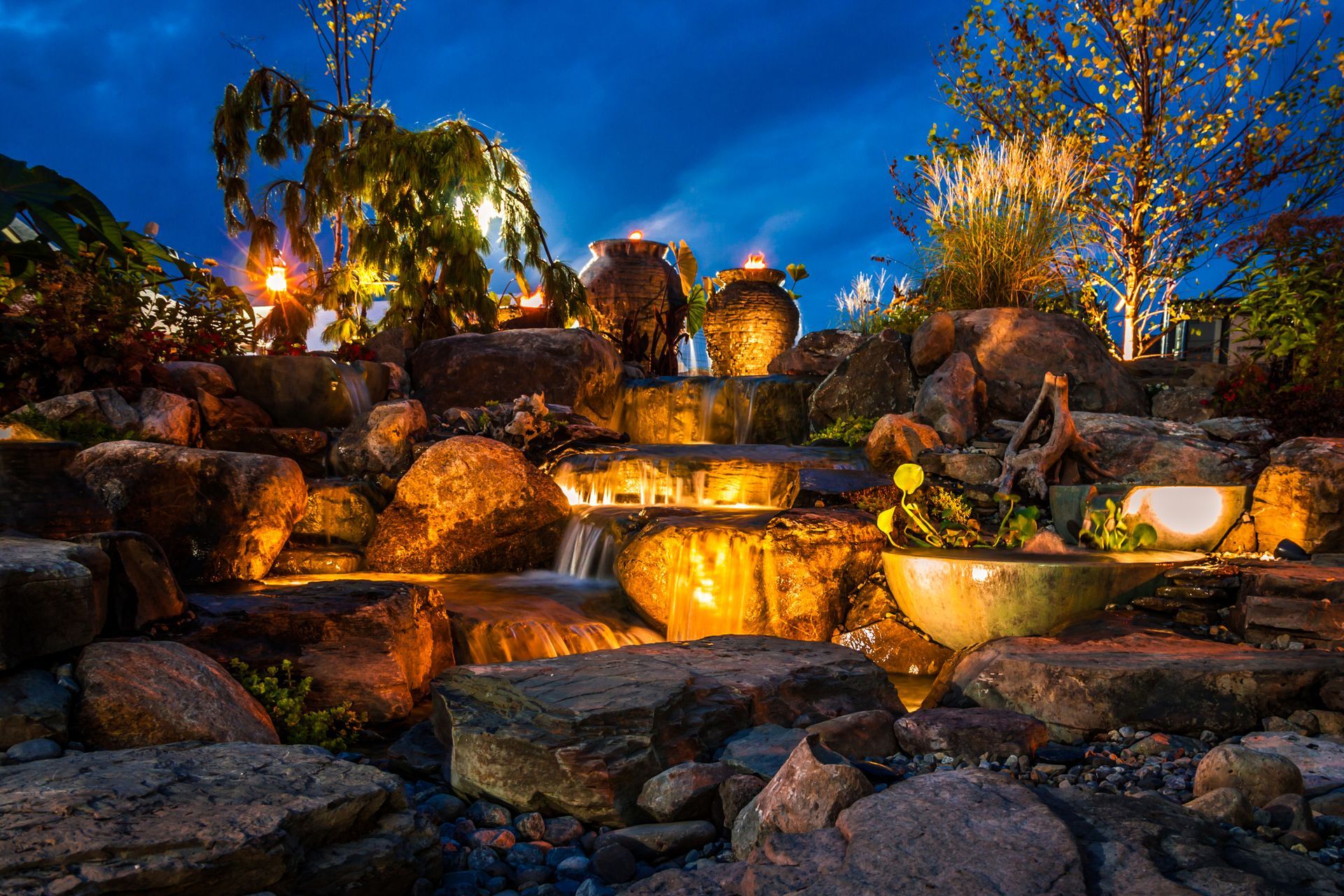 A waterfall surrounded by rocks and trees is lit up at night.